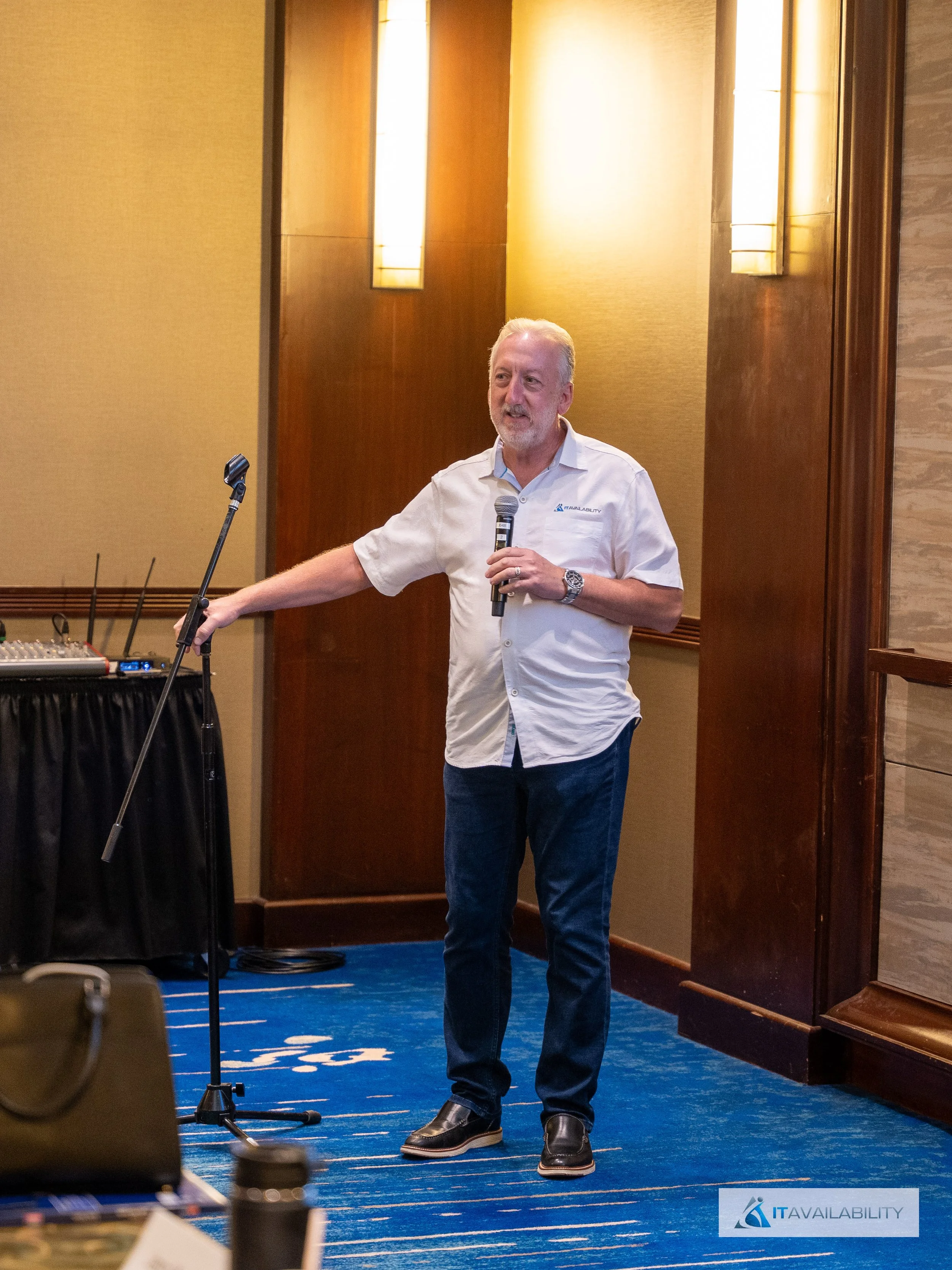 Man giving a presentation or speech with a microphone in hand, standing next to a microphone stand in a conference room with warm lighting and blue carpet.