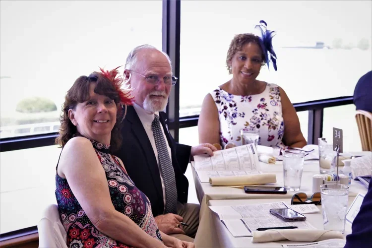 Three adults sitting at a table during a formal event, with two women wearing floral dresses and a man in a suit, all smiling and looking at the camera.