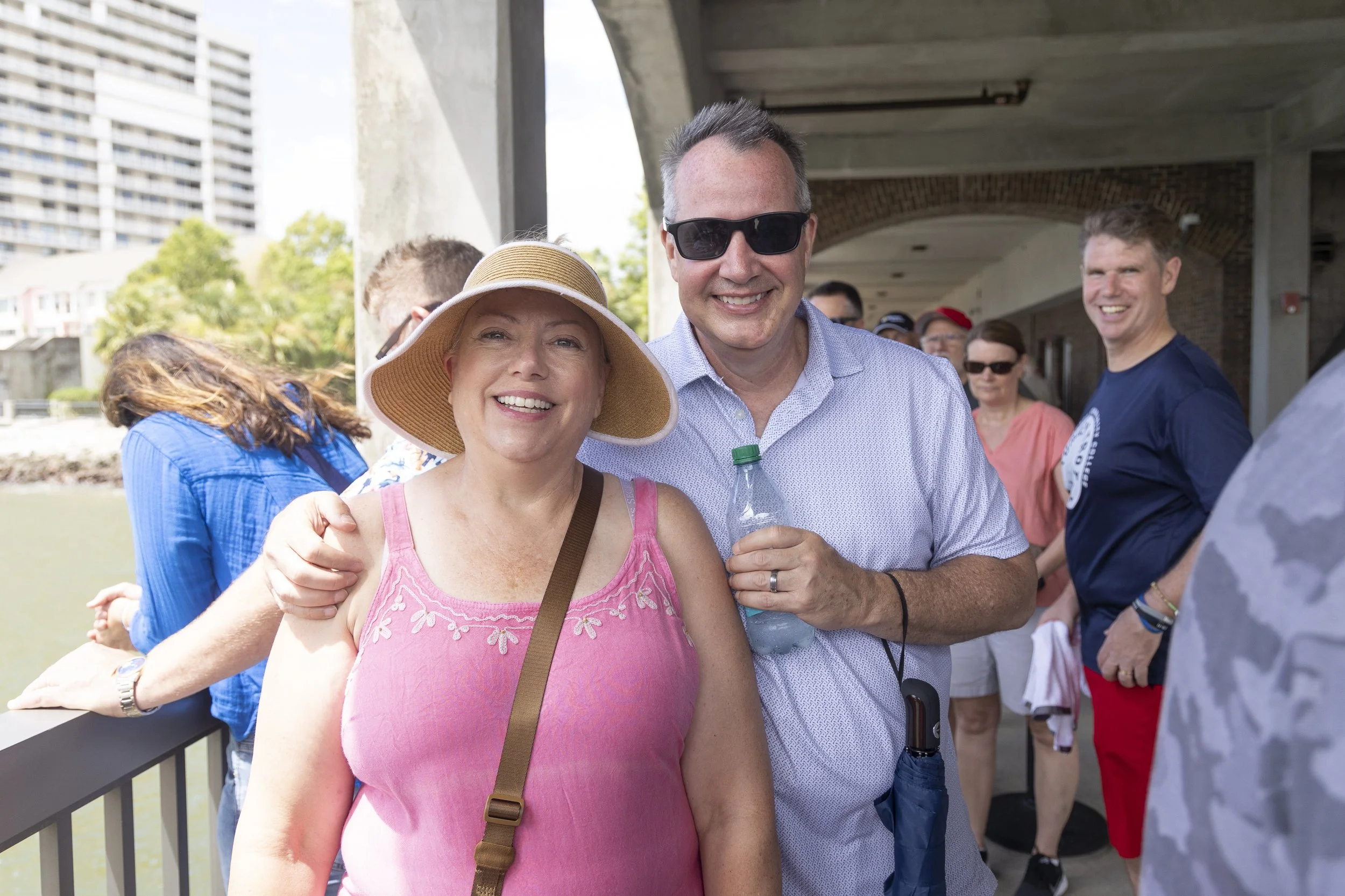 Smiling woman wearing a pink top and wide-brimmed hat standing next to a smiling man in sunglasses holding a water bottle, with a group of people in the background outdoors near a waterfront.