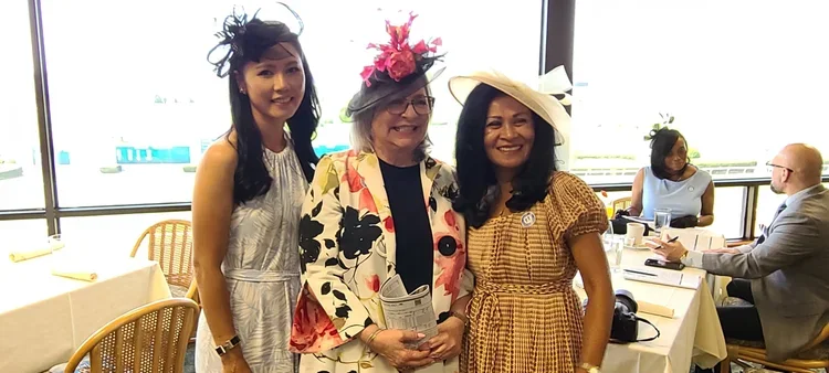 Three women standing together in a room with large windows, dressed in colorful outfits and hats, smiling for the photo. Two other people are seated at a table in the background.