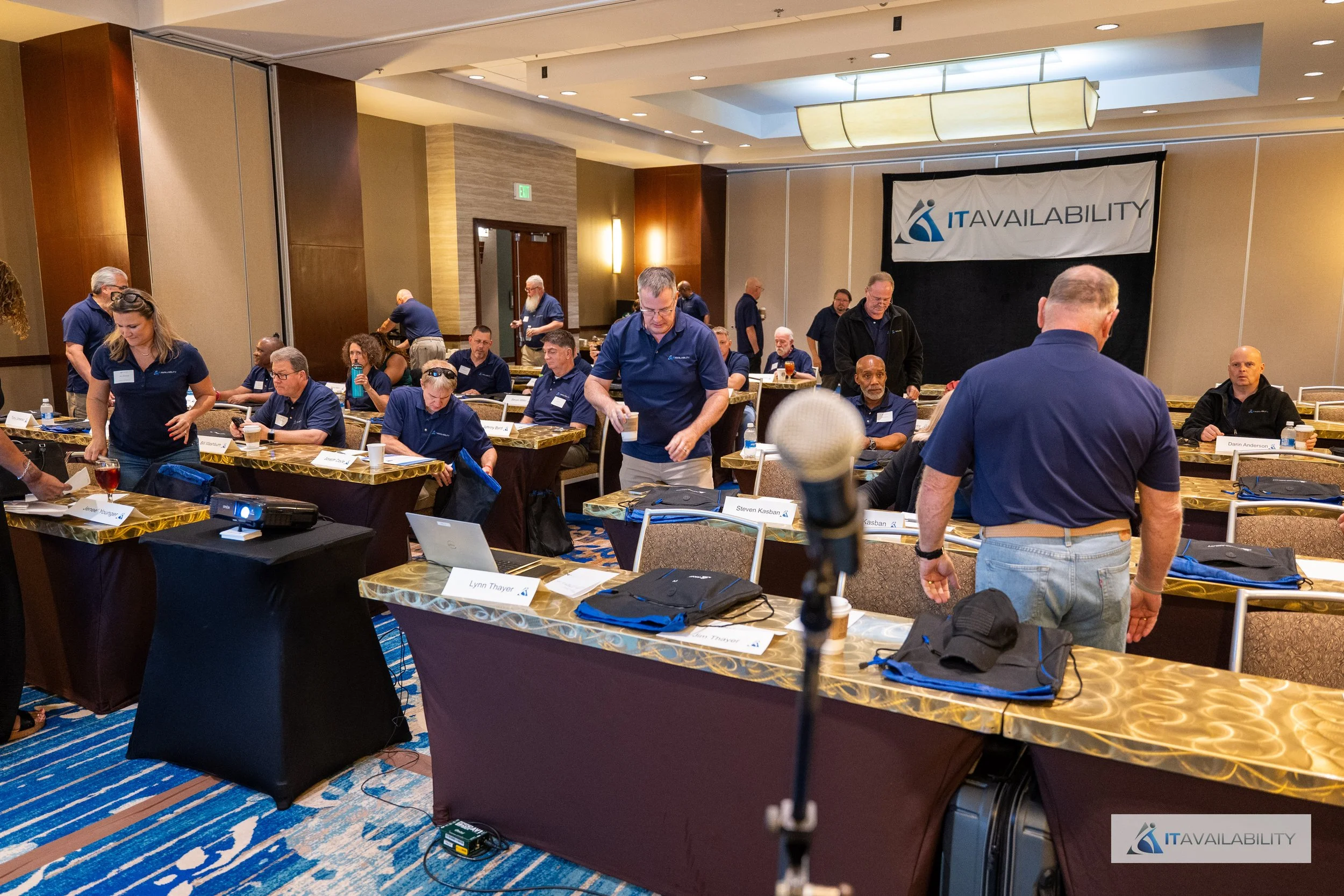 Conference room with people wearing blue shirts, sitting at tables with laptops and papers, preparing for a presentation, with a banner reading 'ITAVAILABILITY' on the wall.