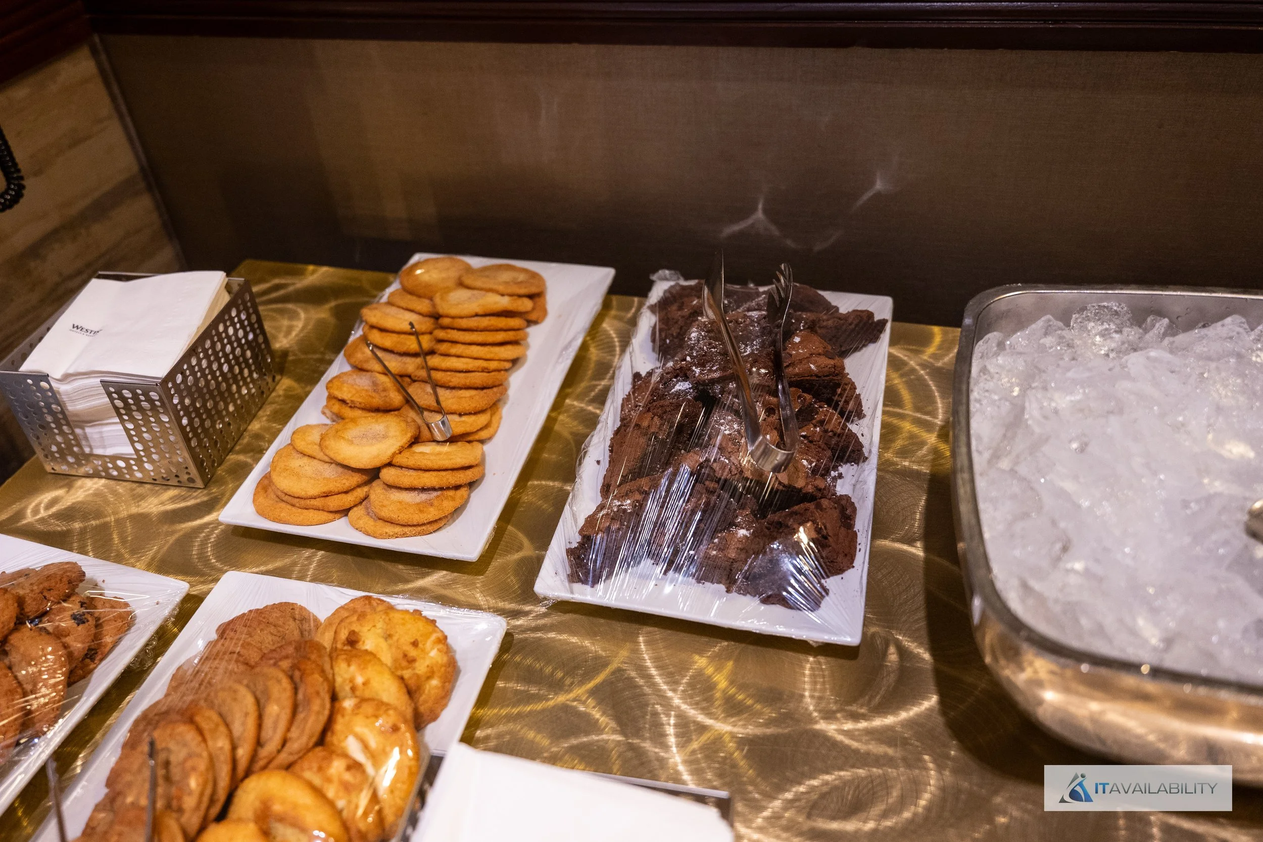 Assorted cookies and brownies on white rectangular platters with a large bowl of ice and beverage service materials on a gold-colored table.