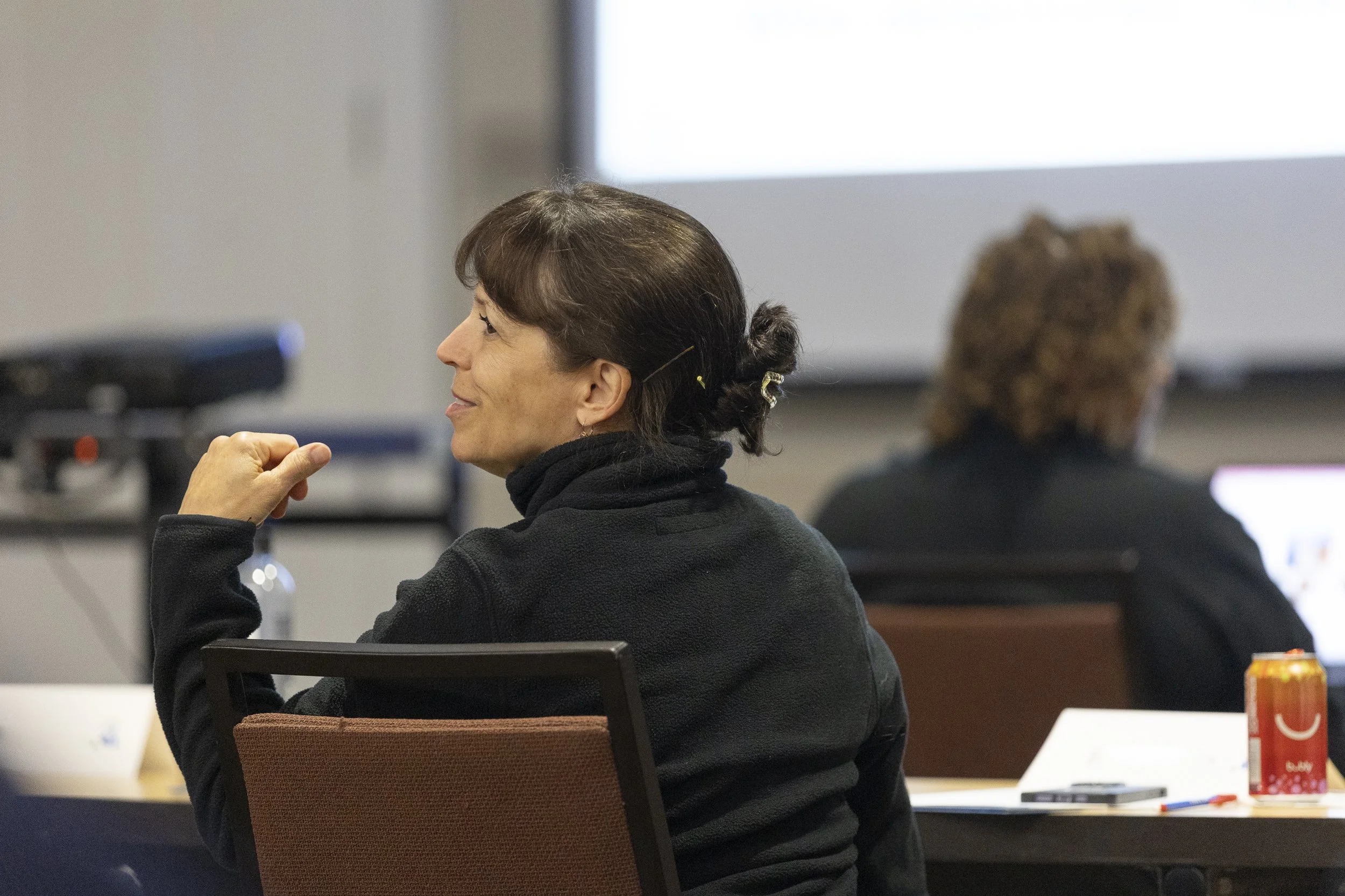 A woman with brown hair in a bun, wearing a black jacket, is sitting at a table in a conference room, smiling and looking to the side. In front of her are papers, a pen, and an orange beverage can. In the background, another person with curly hair is