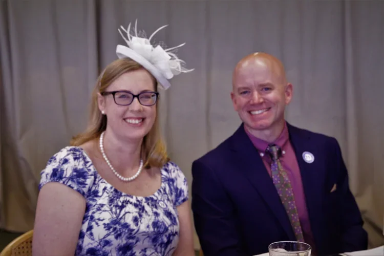 A woman in a floral dress and pearl necklace wearing a white fascinator and glasses, sitting next to a man in a suit, both smiling at a formal event with a gray curtain background.
