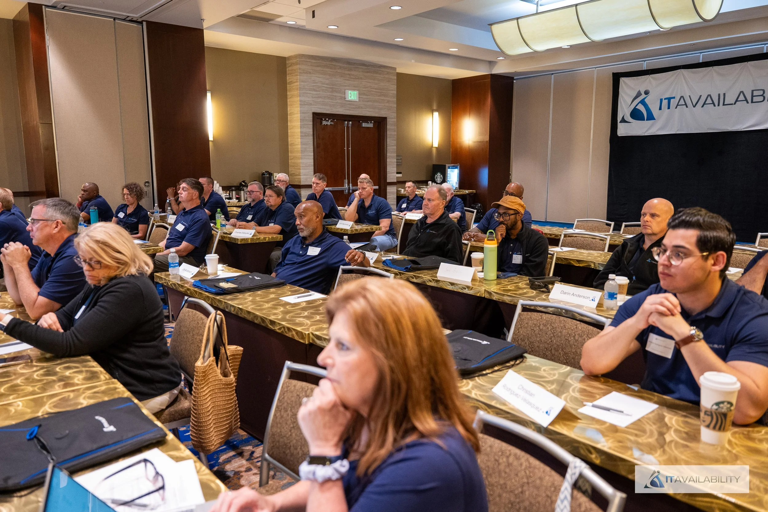 Attendees sitting and listening at a conference in a large hotel ballroom, with tables, notebooks, water bottles, and coffee cups, and a banner that reads "IT Available" in the background.