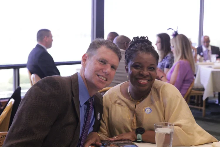 Two smiling people, a man and a woman, sitting at a table during a social gathering or event, with other guests seated at tables in the background.