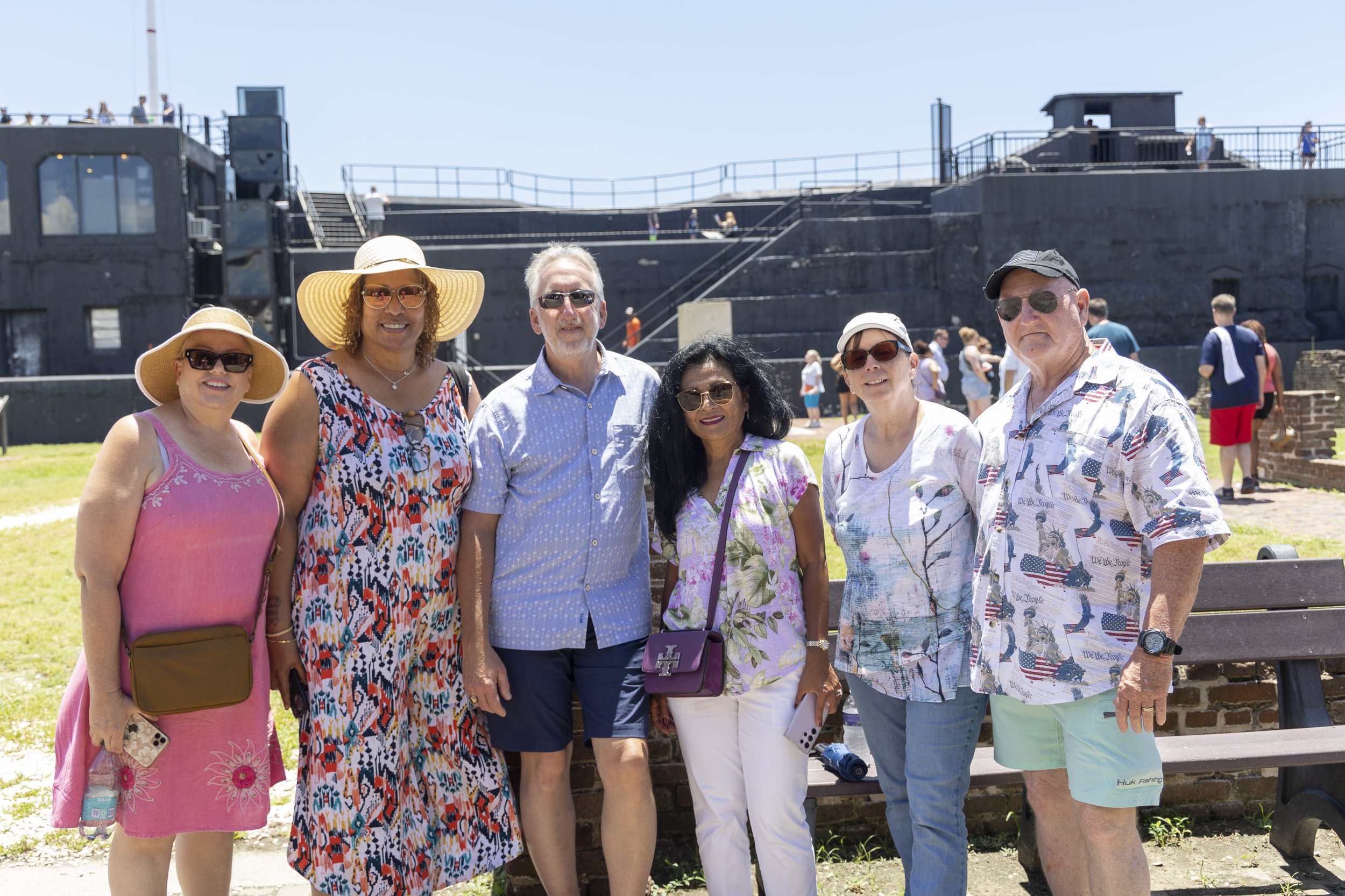 Group of six tourists wearing summer clothing and hats, standing outdoors near a historic black fort on a sunny day.
