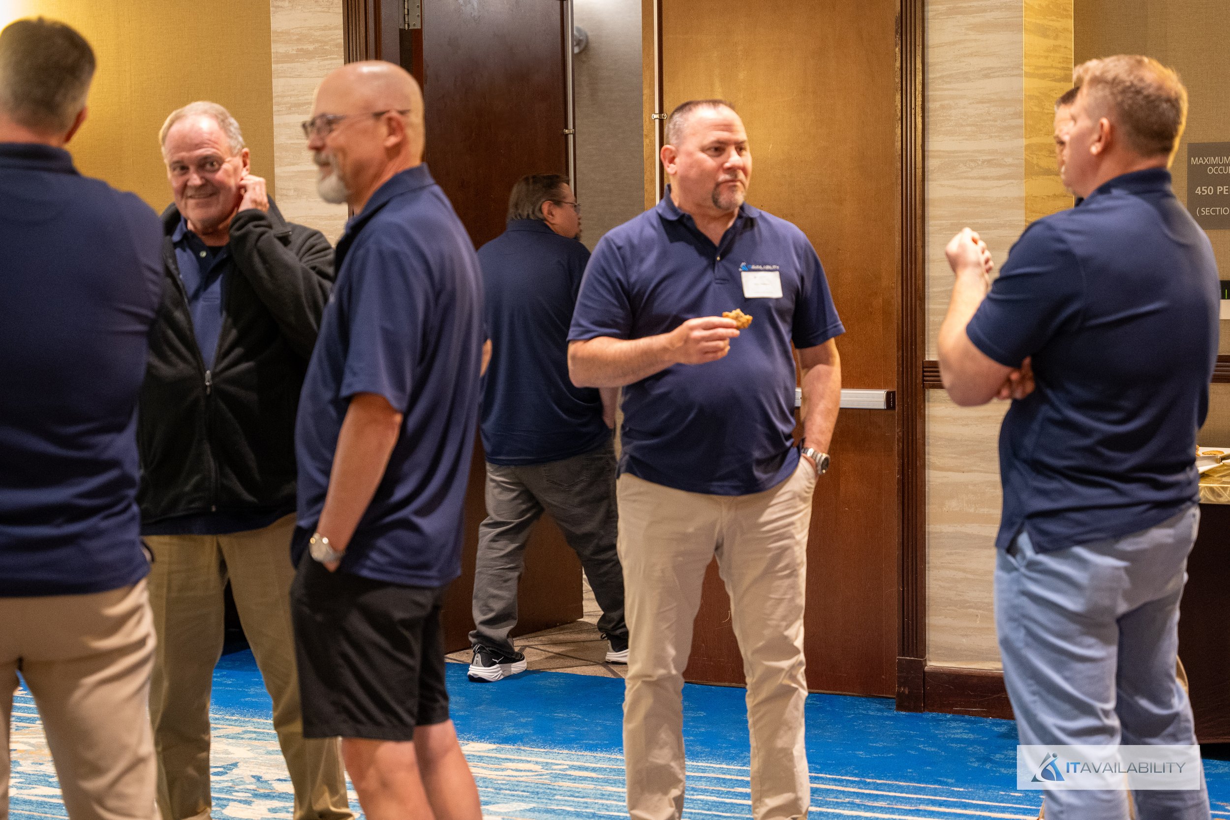 Group of men dressed in navy blue polo shirts and khakis talking at a conference or networking event in a hotel hallway.