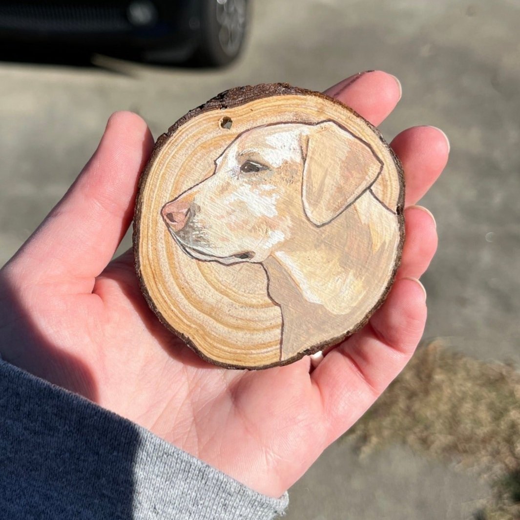 A hand holding a wooden slice with a painting of a dog's profile on it, outdoors with a blurred background.
