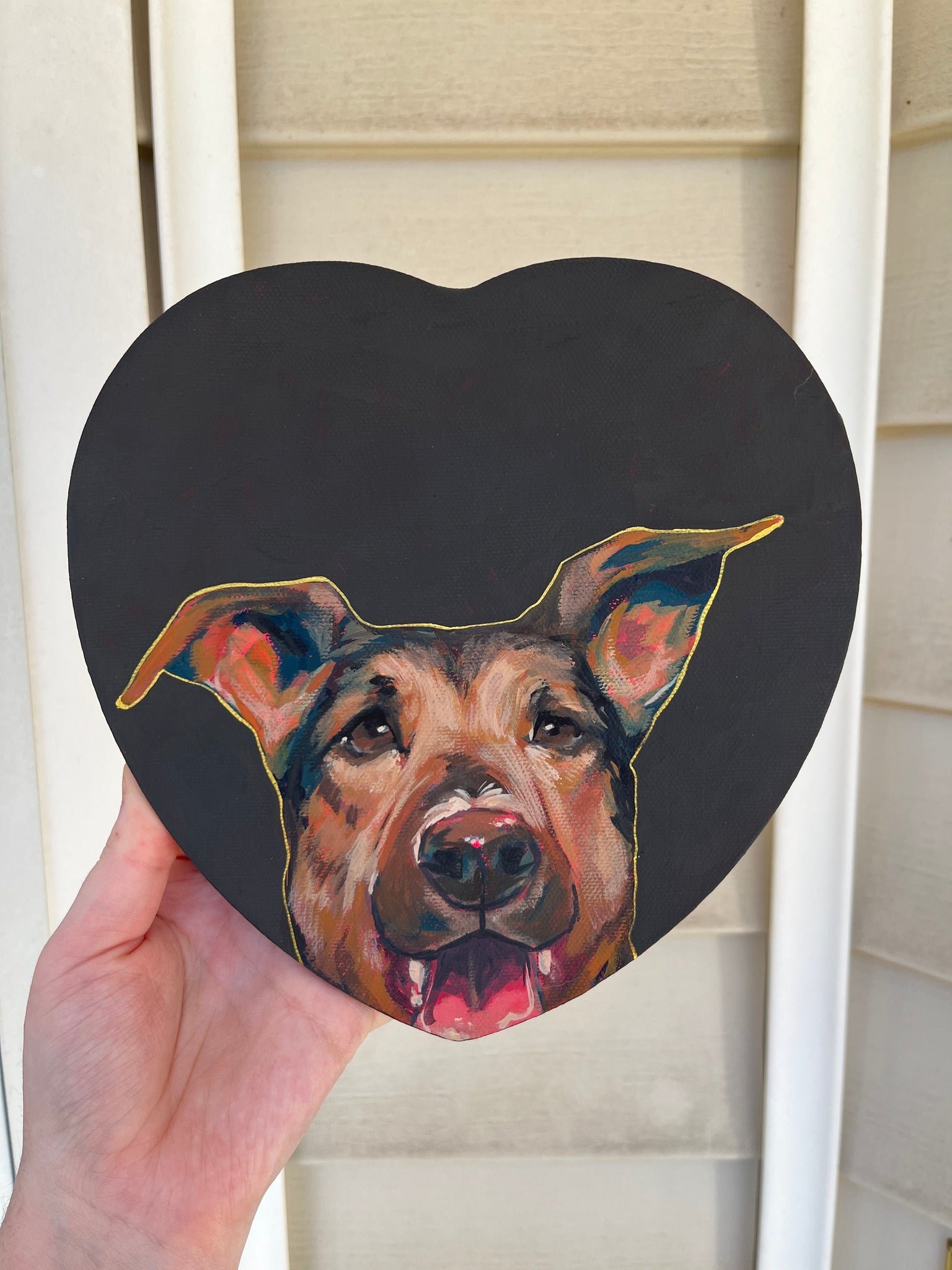 A black heart-shaped sign with a colorful painted portrait of a happy, smiling dog with its tongue out.