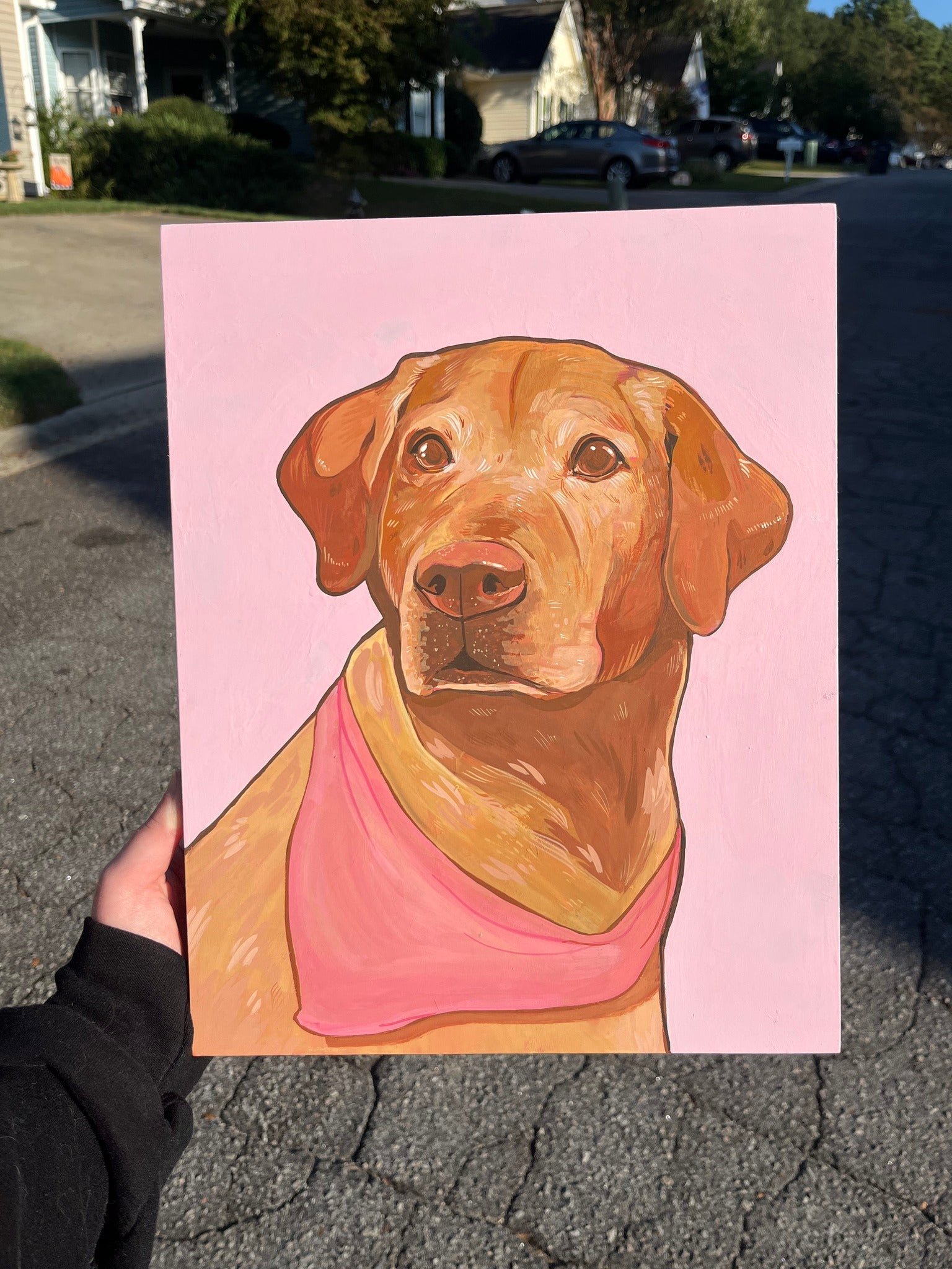 Painting of a brown dog with a pink bandana on a pink background, held in front of a residential street.