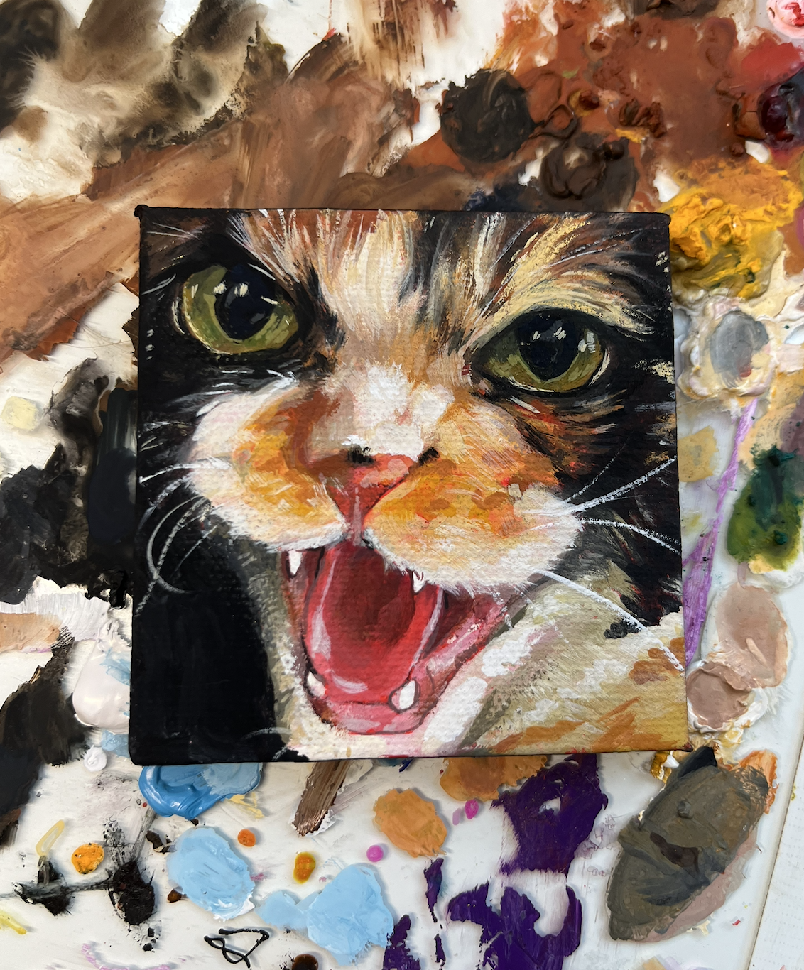 A painting of a cat with yellow-green eyes and an open mouth, showing its tongue and teeth, with a messy palette of oil paints around it.