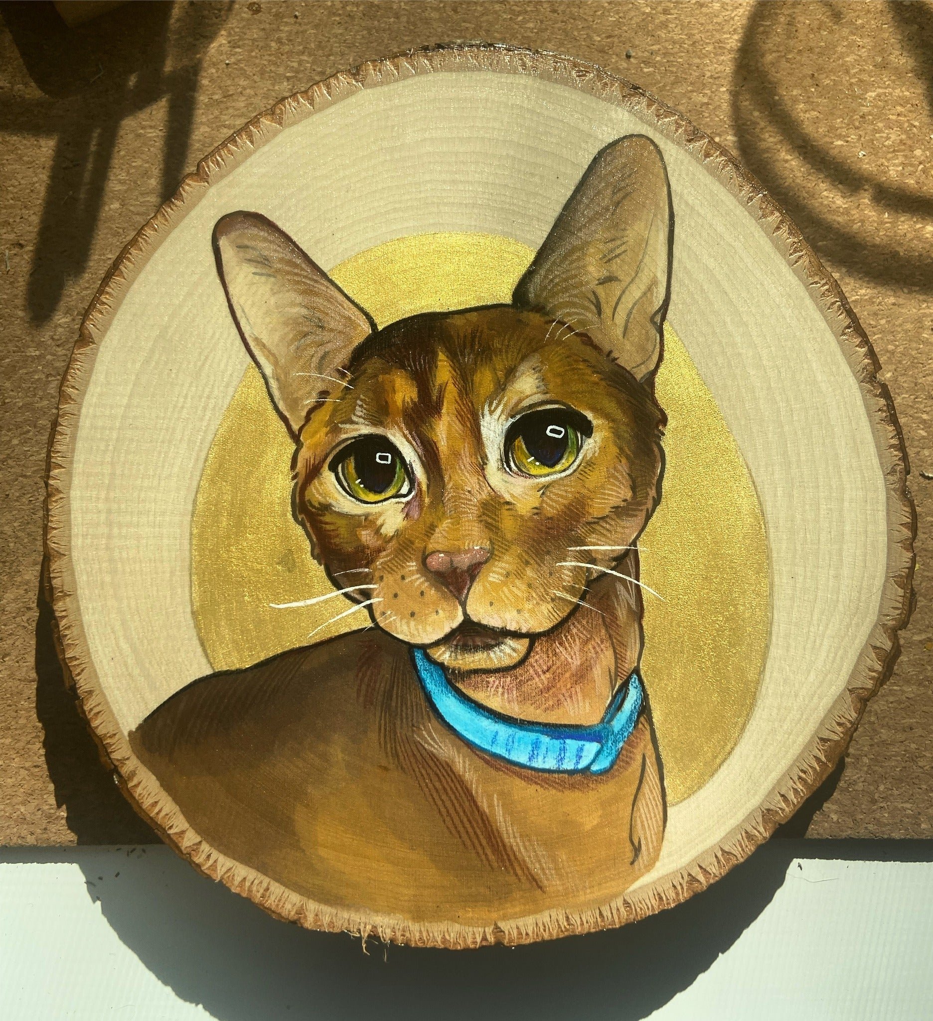 Painted portrait of a brown cat with green eyes and a blue collar on a wooden slice background.