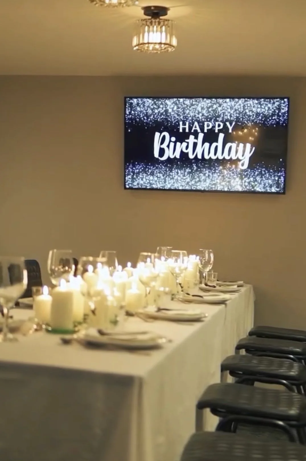 A decorated birthday celebration with a long table set with plates, glasses, and lit candles, in front of a digital screen displaying a 'Happy Birthday' message with a sparkly background.