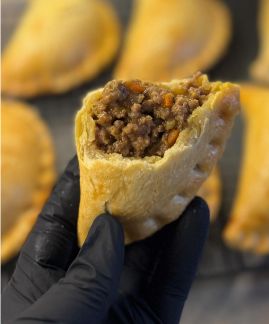 Close-up of a hand in a black glove holding a partially eaten beef and vegetable potstickper with a golden-brown exterior, with more potstickers in the background.