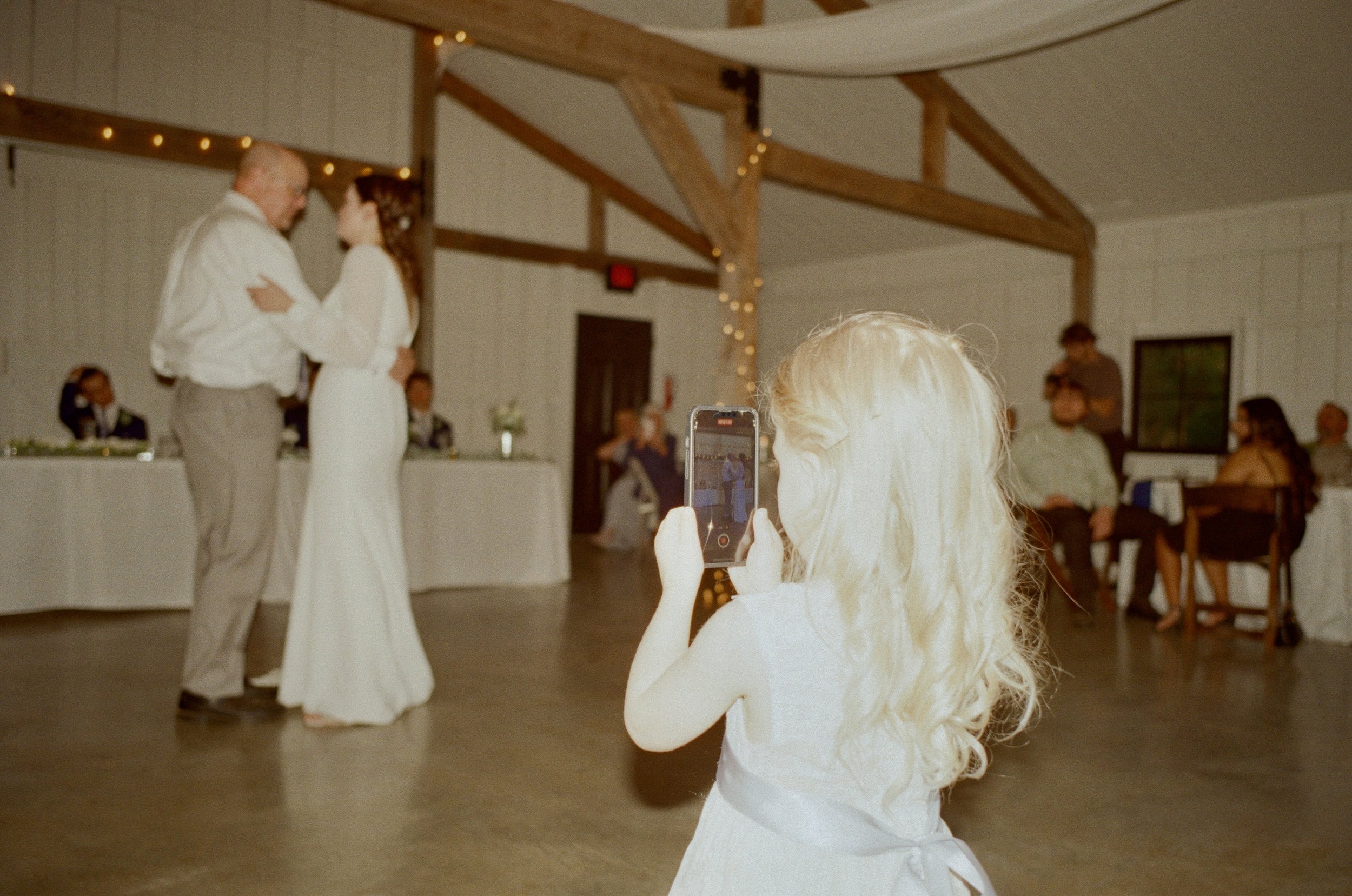 A young girl with blonde hair wearing a white dress is taking a photo of a couple dancing at a wedding reception. The bride and an older man are embracing and dancing in front of a decorated table and guests seated at tables in a venue with wooden be