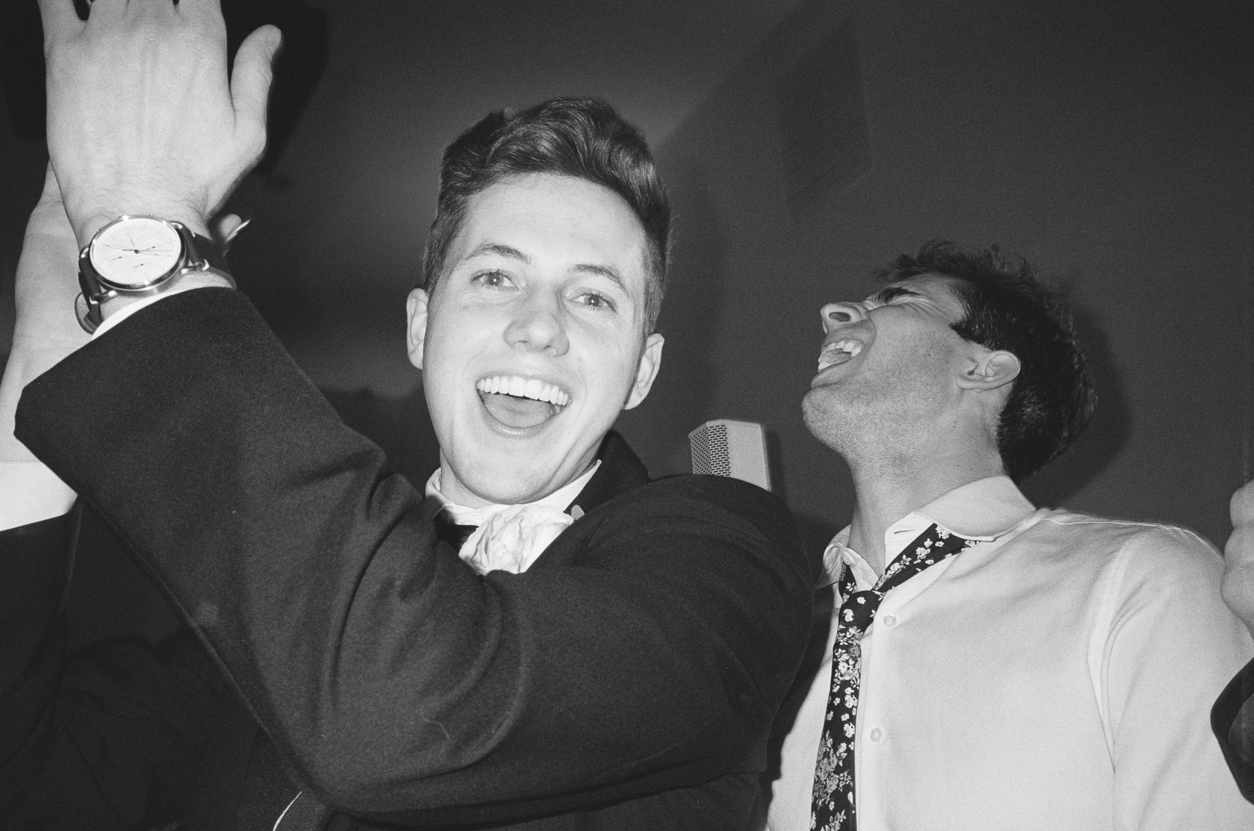 Two men celebrating and laughing, with one wearing a suit and tie, in a joyful moment.