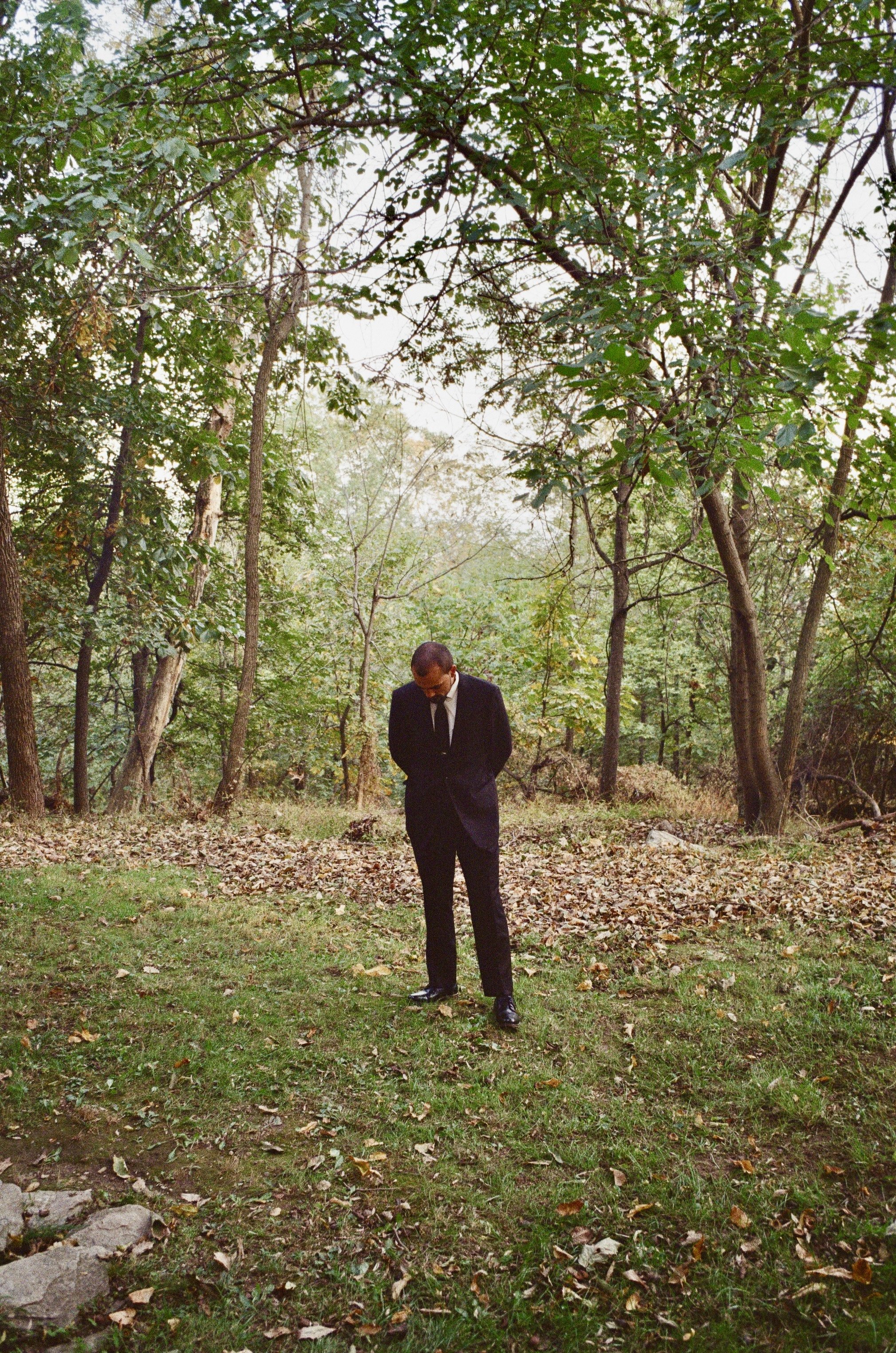 A man dressed in a black suit and tie walking alone in a wooded area with green trees and fallen leaves.