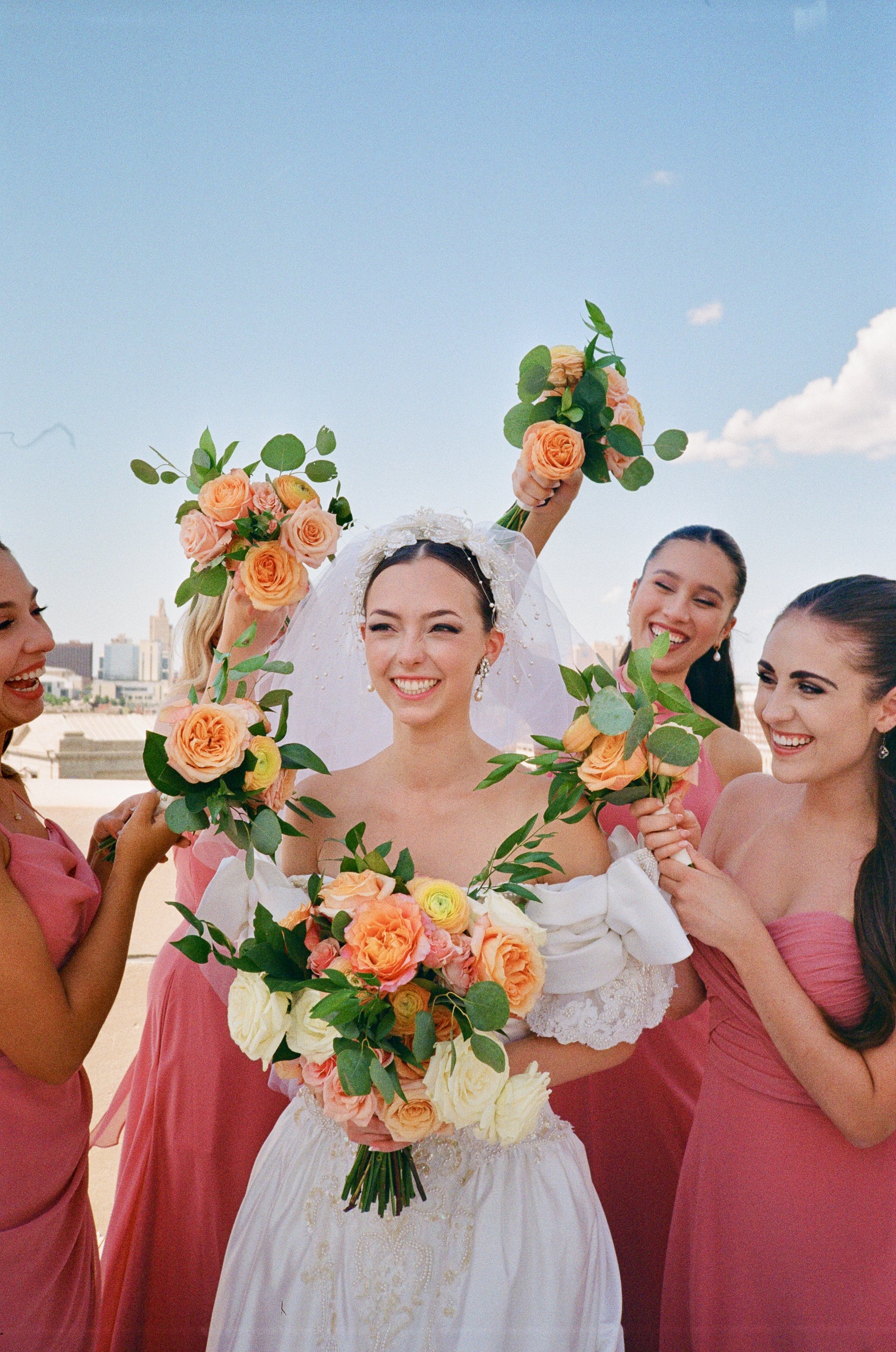 A bride in a white wedding dress smiling and holding a large bouquet, surrounded by four bridesmaids in pink dresses, all holding smaller bouquets, on a rooftop against a blue sky with a city skyline in the background.