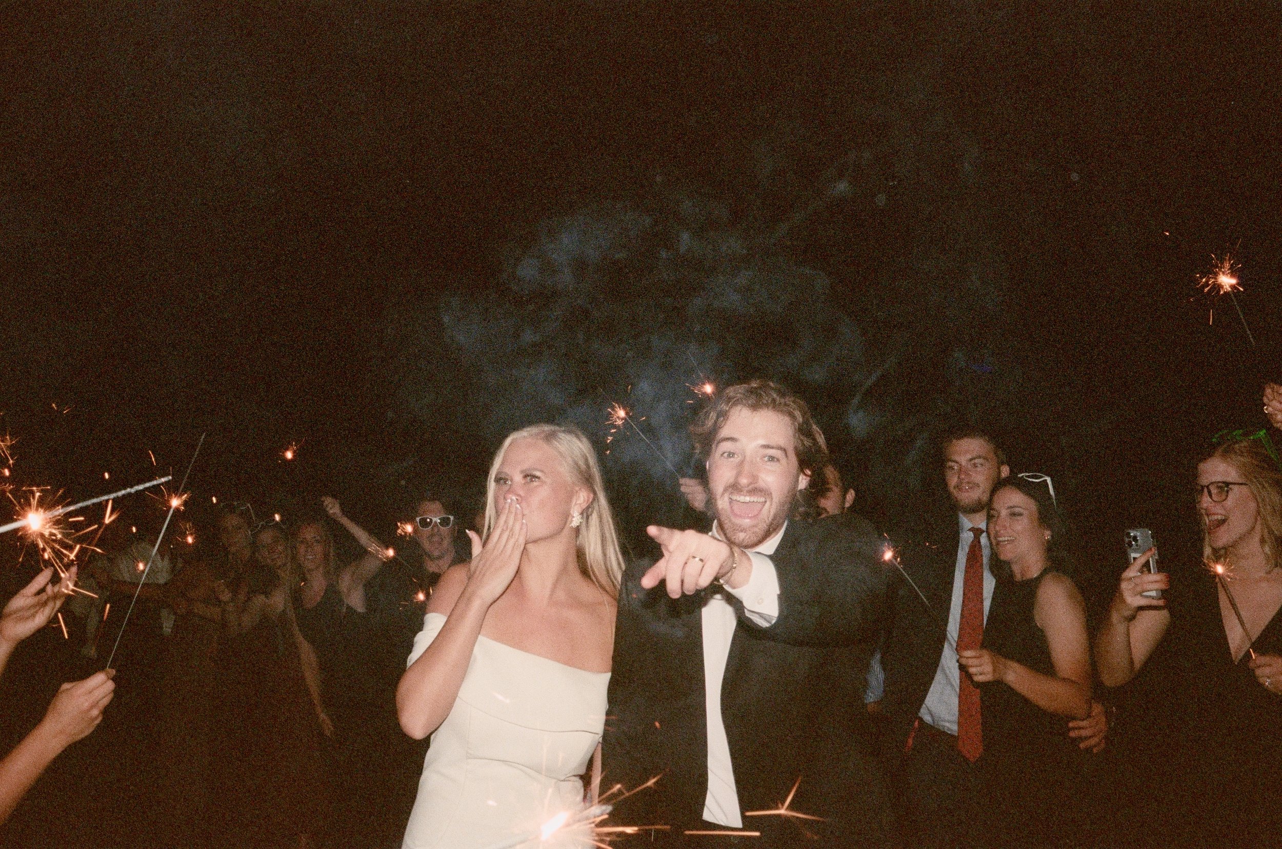 People celebrating at night with sparklers, dressed in formal attire, smiling and enjoying a festive event.