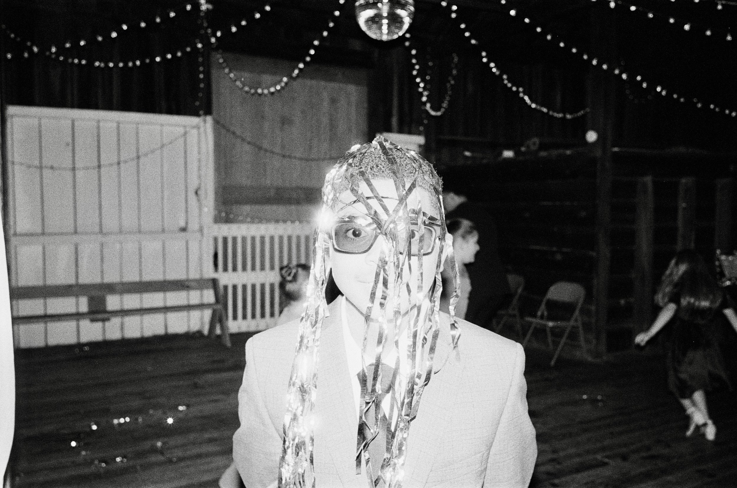 Person in a suit with a mask covered in metallic streamers at a party with string lights and children dancing in the background.