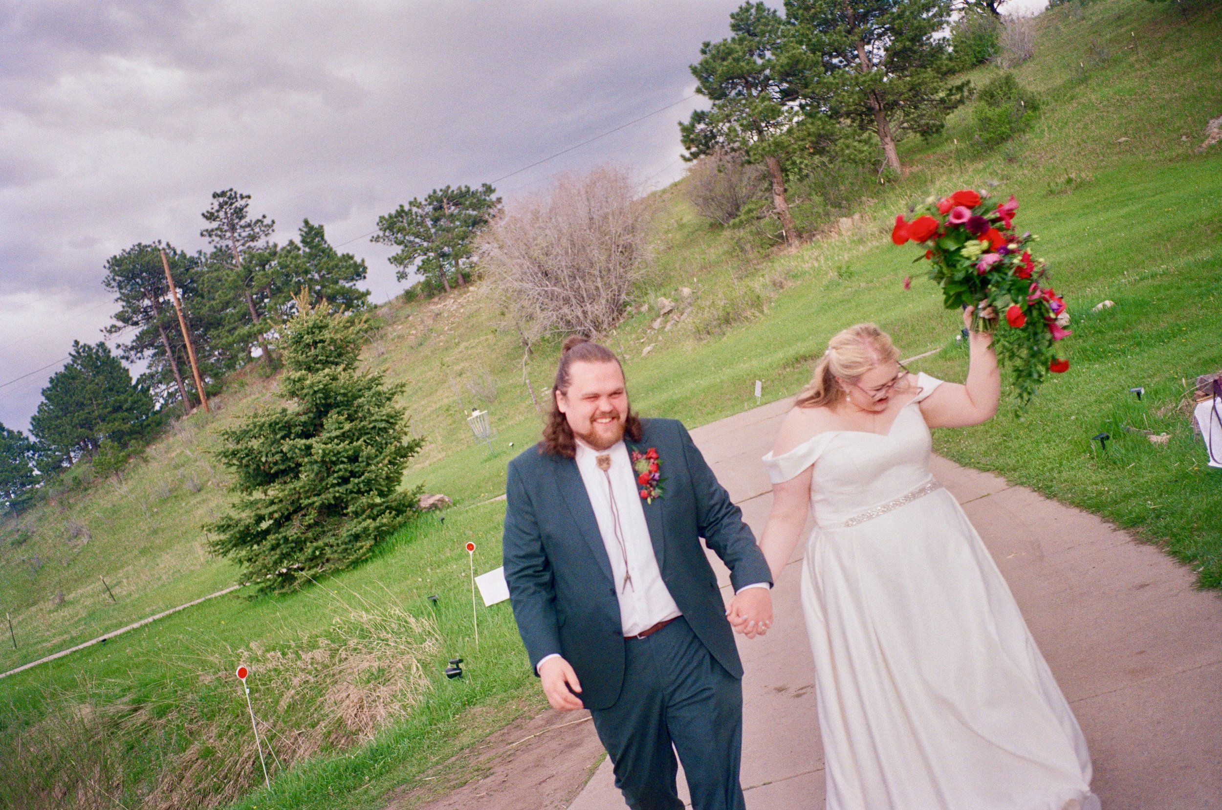 A newlywed couple walking outdoors on a cloudy day, holding hands with the bride carrying a large bouquet of red and pink flowers.