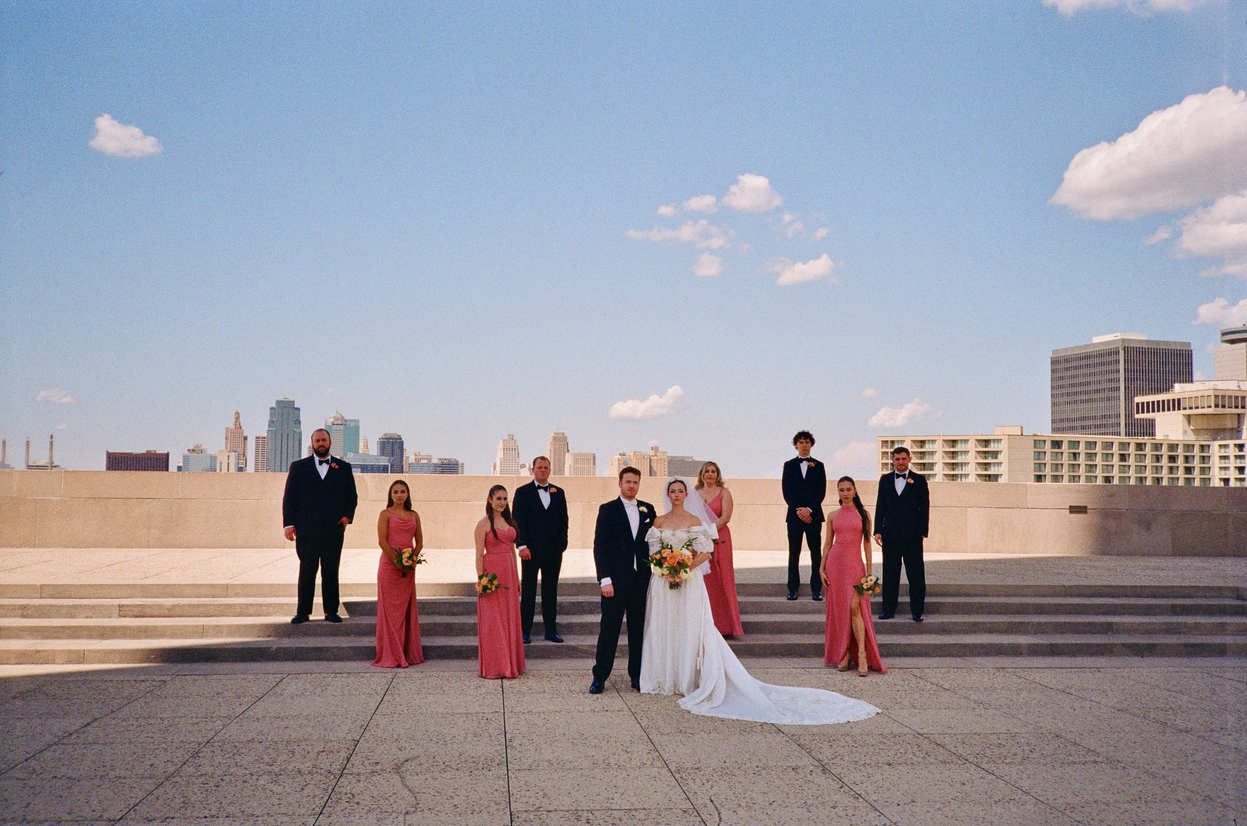 A wedding party on a city rooftop with a city skyline in the background, including a bride in a white gown and seven bridesmaids and groomsmen in formal attire.