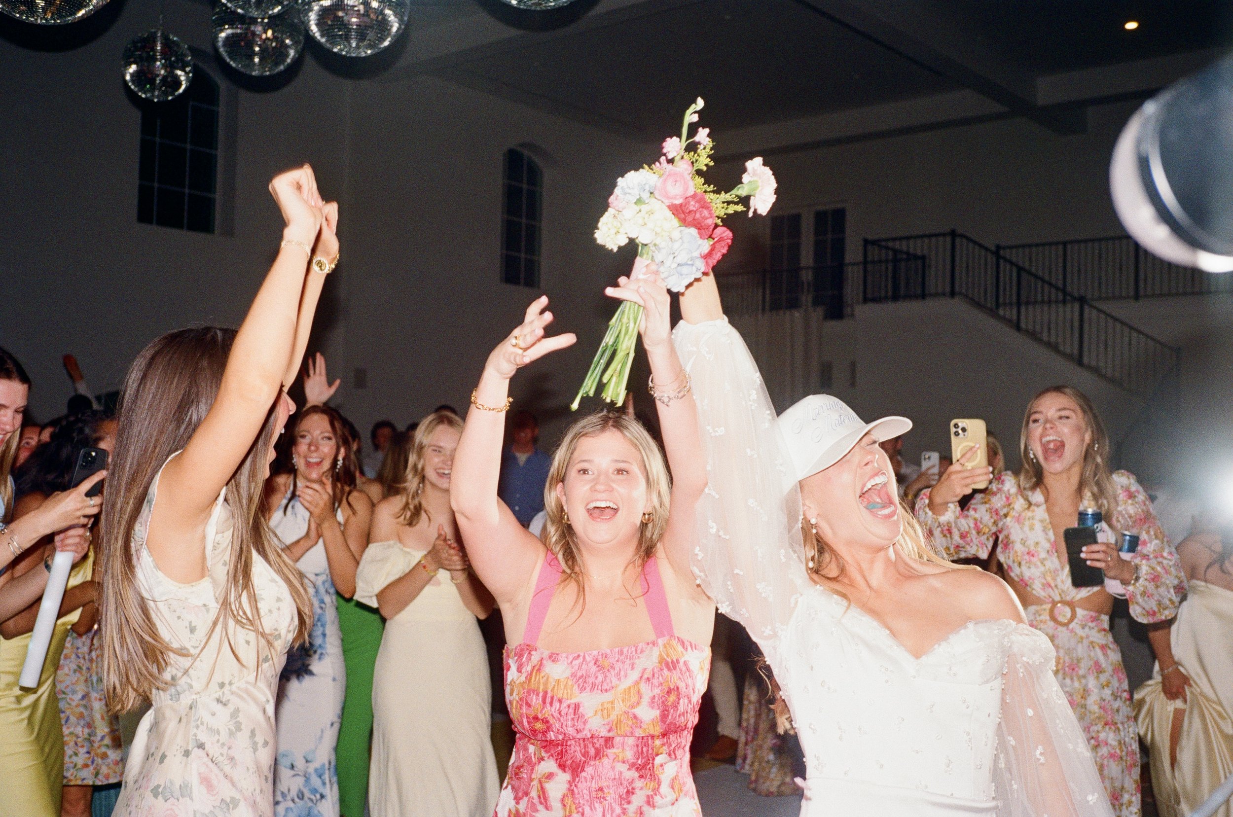 A woman in a wedding dress holding a bouquet celebrates with friends at a wedding reception. The friends are smiling and taking pictures.