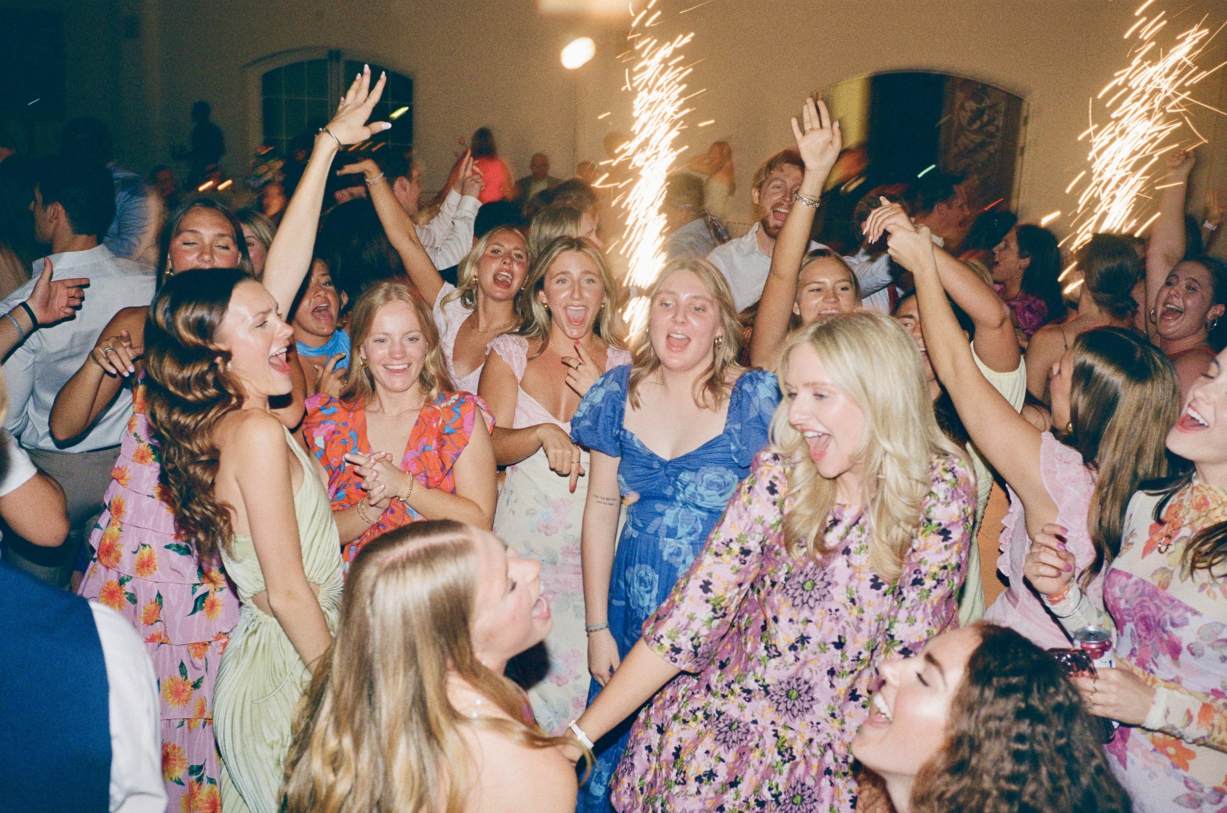 A lively party scene with a group of young women and men dancing and celebrating, fireworks and sparklers in the background, smiling and laughing.