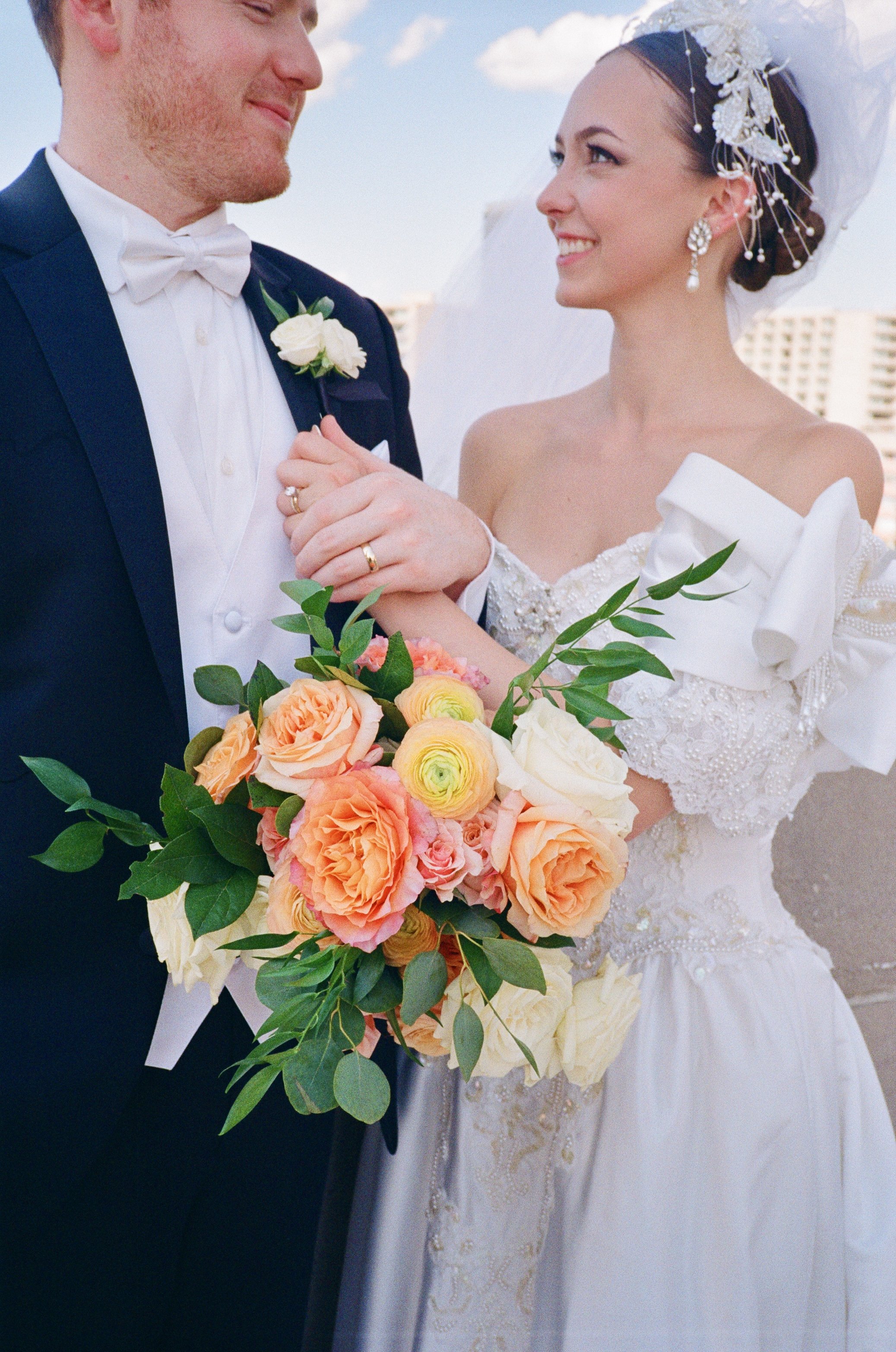 A bride and groom dressed in wedding attire standing together, with the bride holding a bouquet of peach, cream, and pink flowers, on a rooftop with a cityscape in the background.
