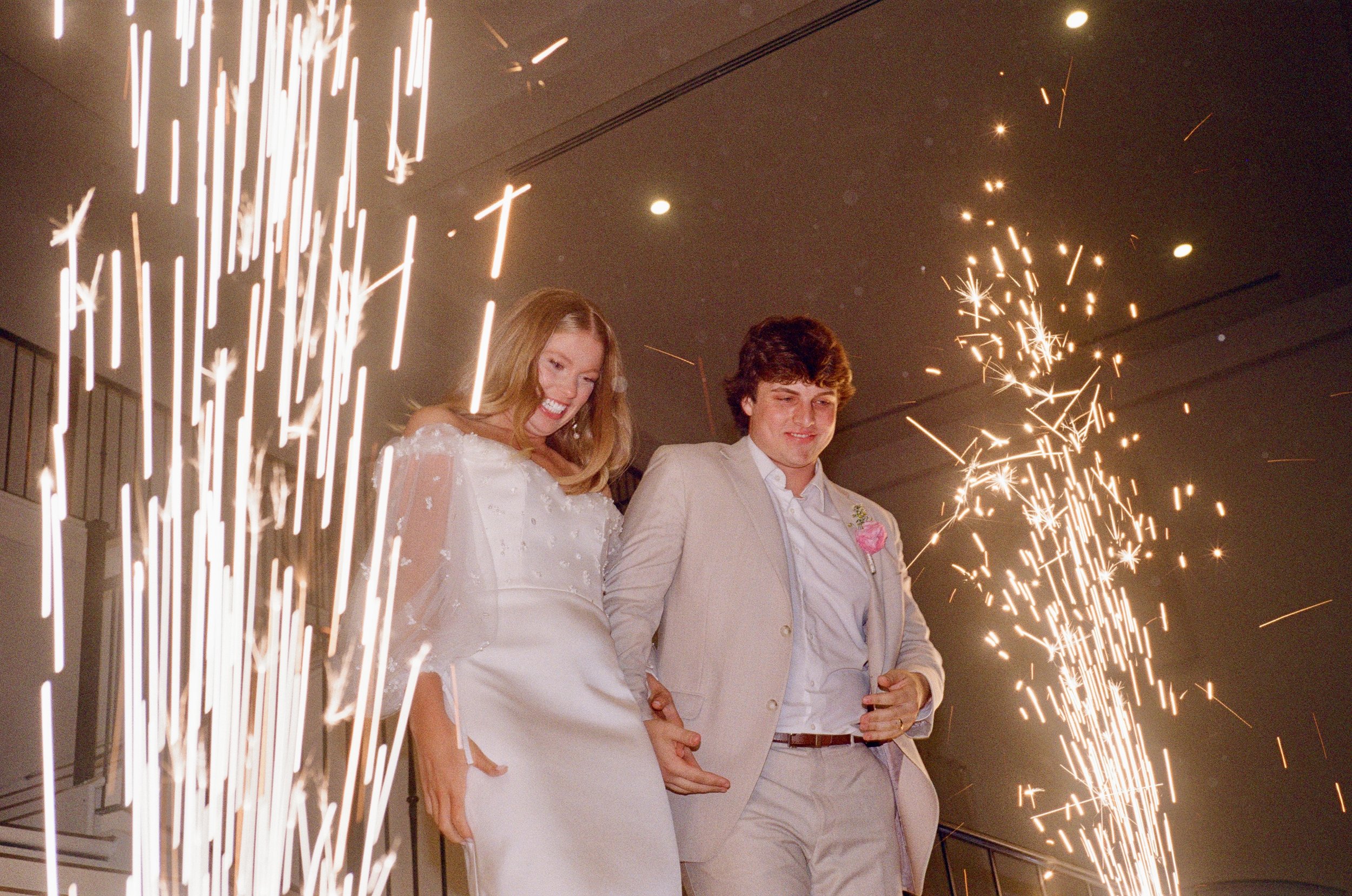 A bride and groom smiling as they walk through sparklers at their wedding celebration.