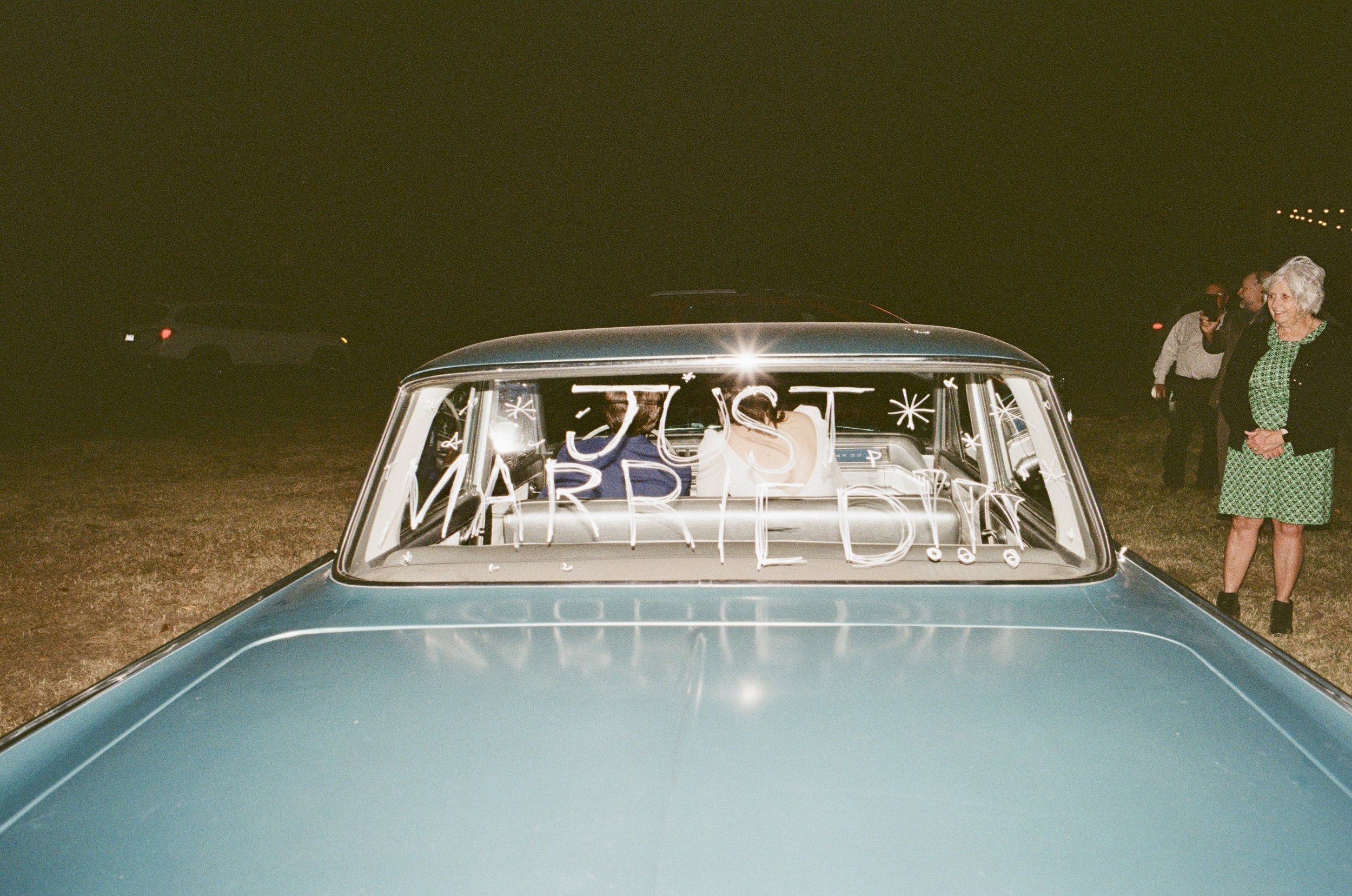 The front of a vintage car with a sign reading 'JUST MARRIED!!' on the windshield, and a couple inside. Two women stand outside the car, one smiling, at night.