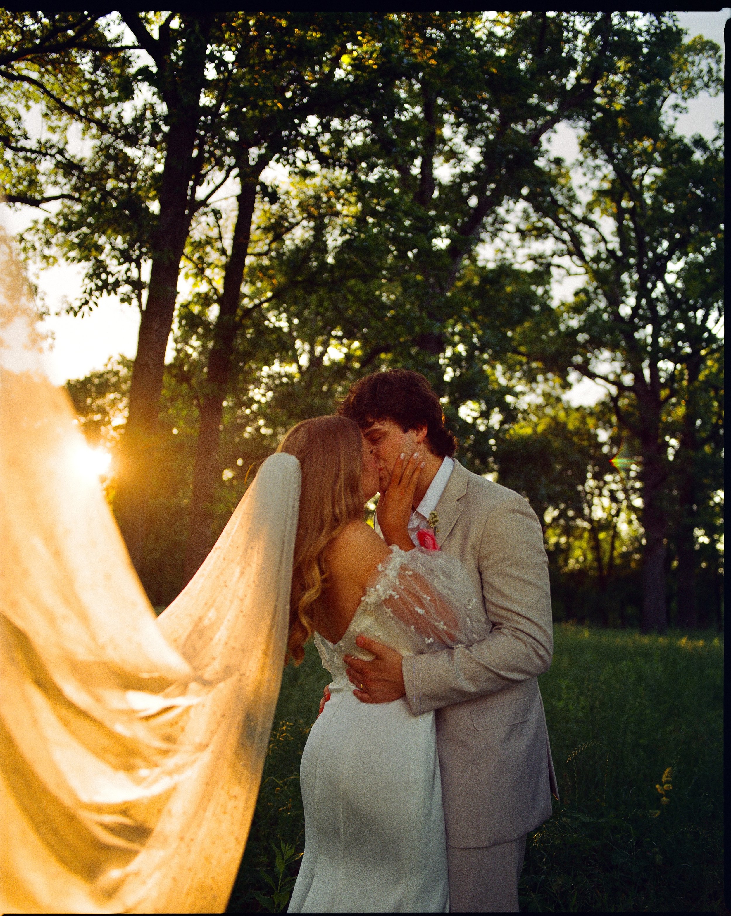 A couple is kissing outdoors during sunset, with trees in the background, on their wedding day. The bride is wearing a long white wedding gown and veil, and the groom is in a light-colored suit.