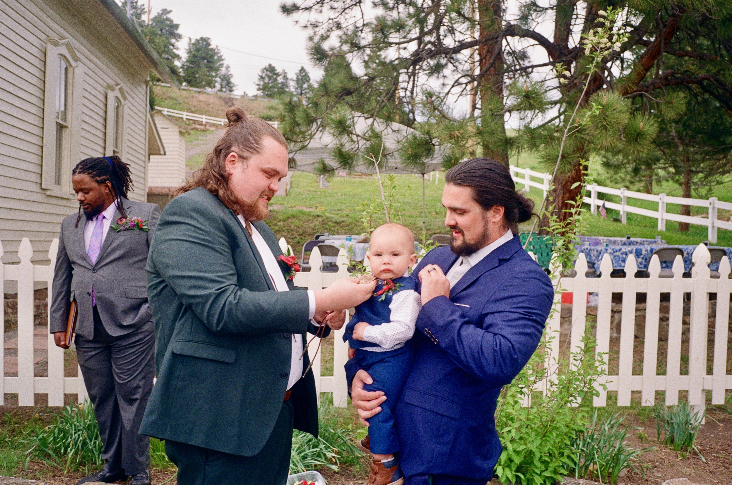 Groom and man holding a baby outside in a garden with white picket fence and trees