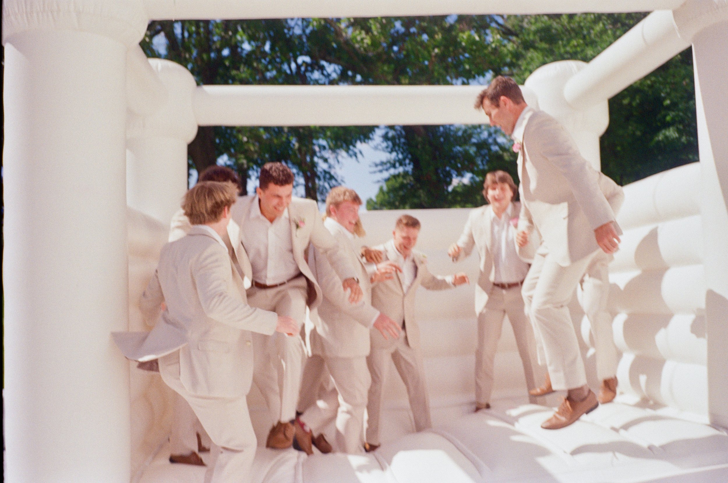 Group of men in light-colored suits and brown shoes having fun on a white inflatable bounce house outdoors on a sunny day.