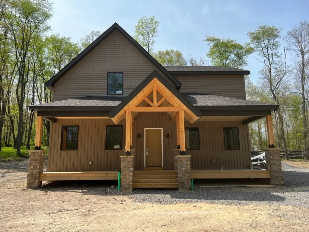 New two-story house under construction with wooden porch and brick columns, set in a wooded area.