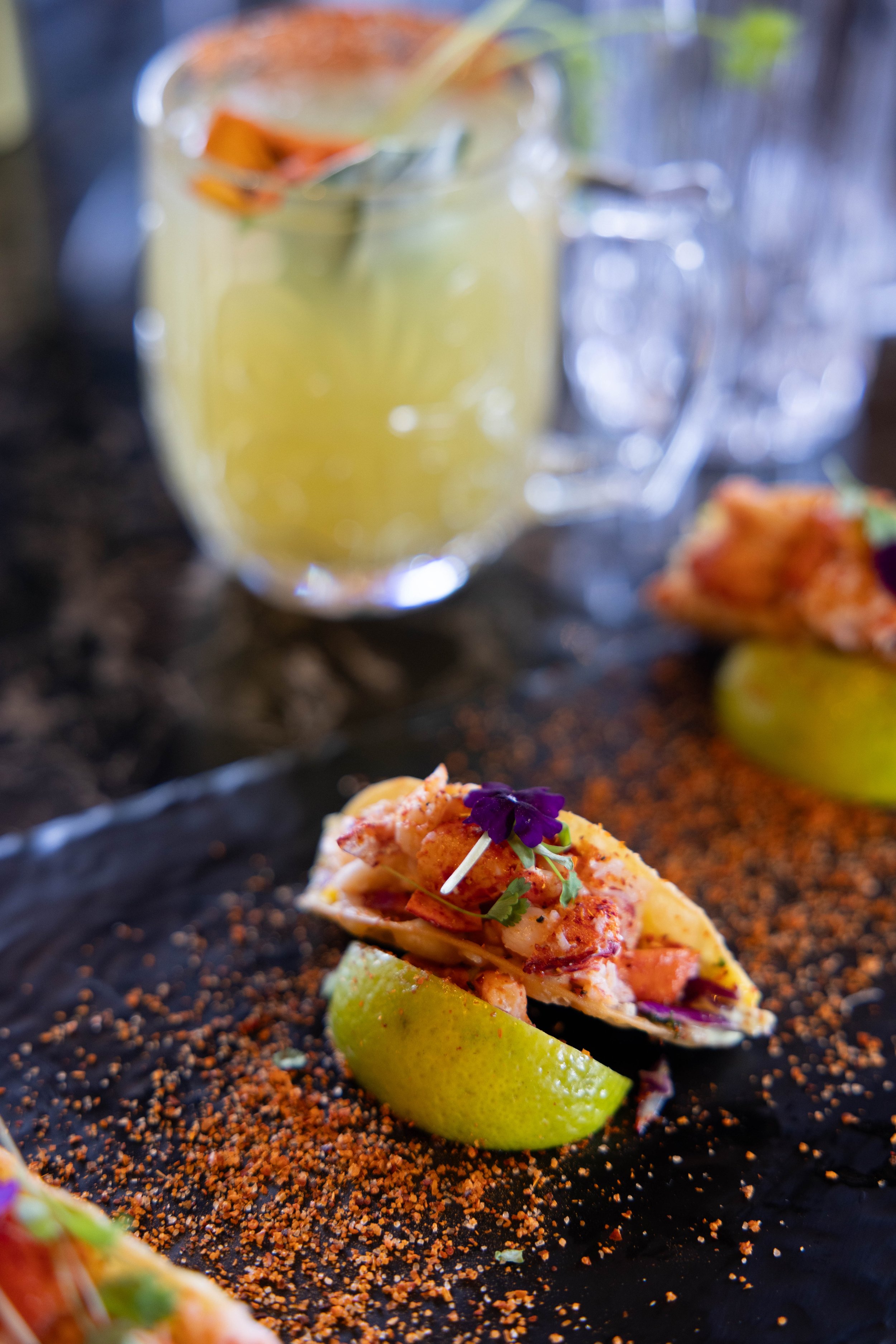 Close-up of a seafood taco on a black plate, garnished with a purple edible flower and cilantro, with a lime wedge underneath, sprinkled with red spice, and a jar of lemon water in the background.