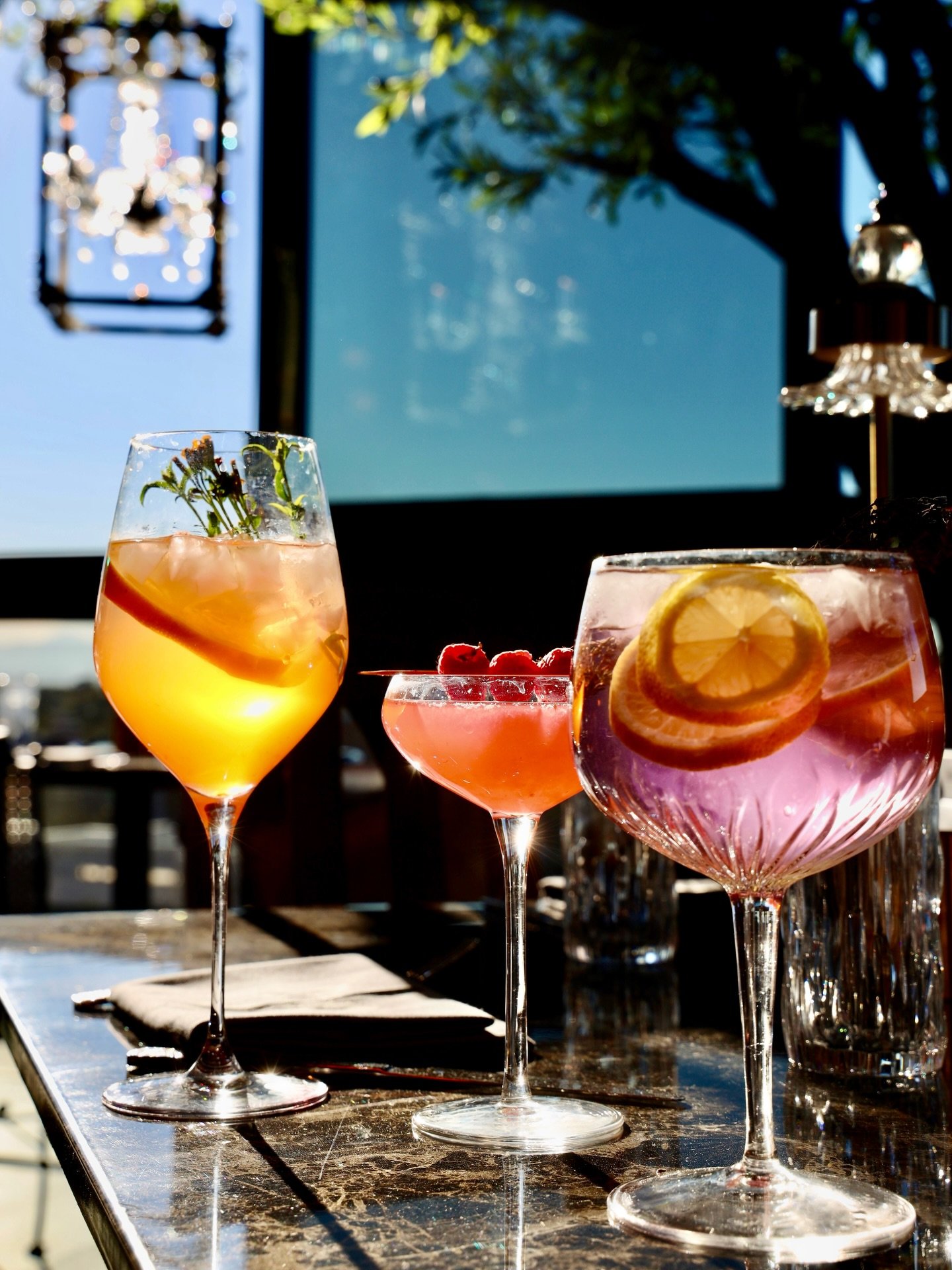 Three colorful cocktails with fruit garnishes on a table outdoors, with sunlight and reflections.