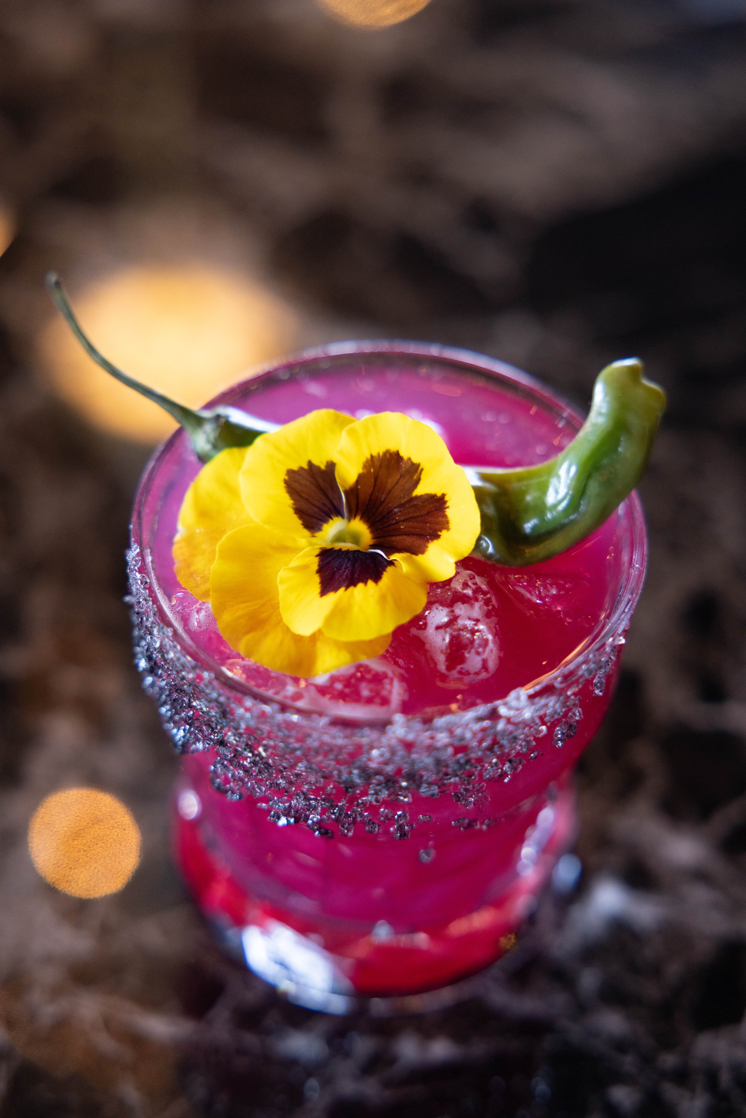 A pink cocktail in a glass with sugar rim, garnished with a yellow and brown pansy flower, a green chili pepper, and ice cubes.