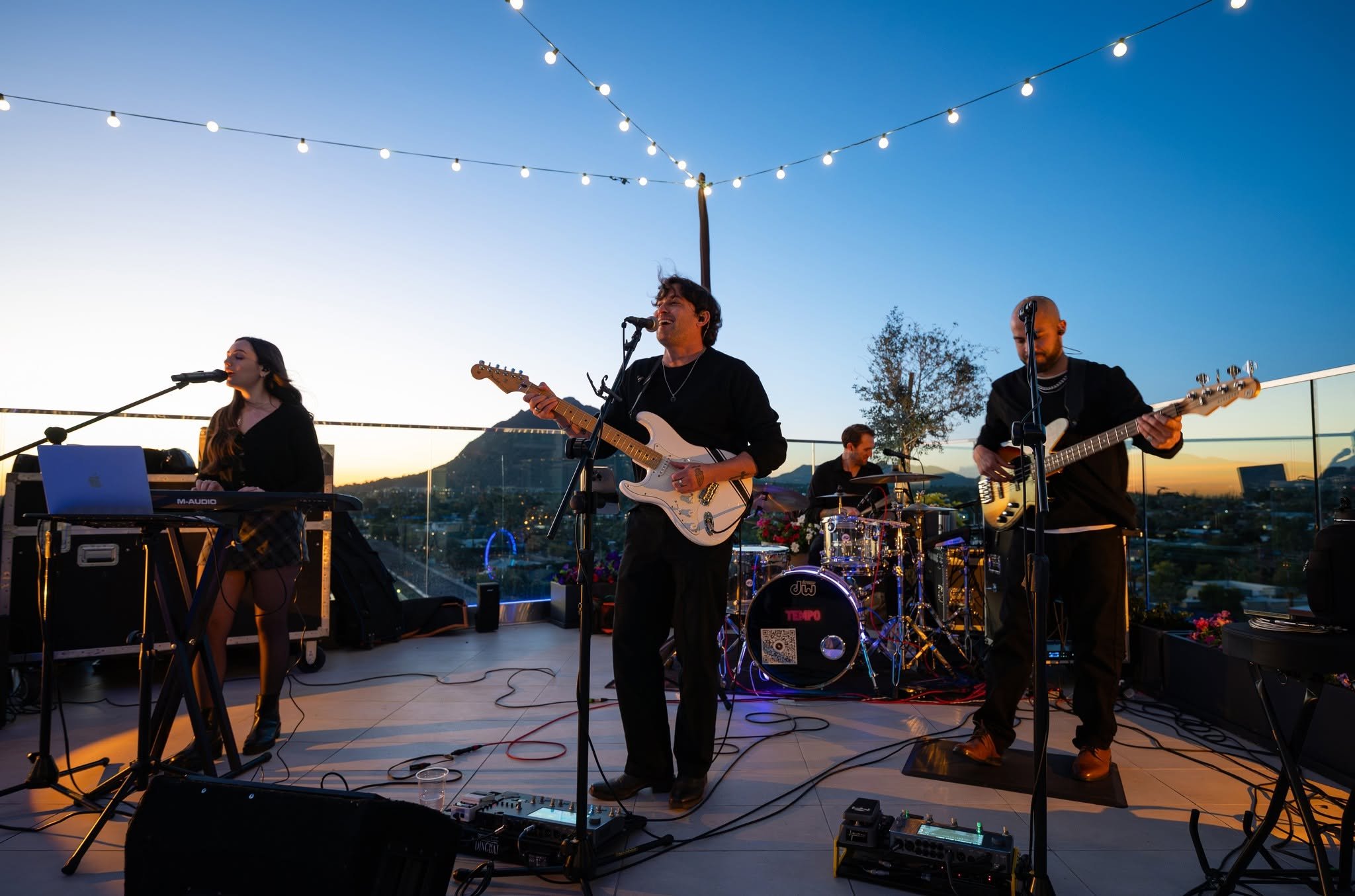 Band performing on an outdoor rooftop stage during sunset, with string lights overhead, cityscape in the background, and various musical instruments including guitars, keyboard, drums, and microphones.