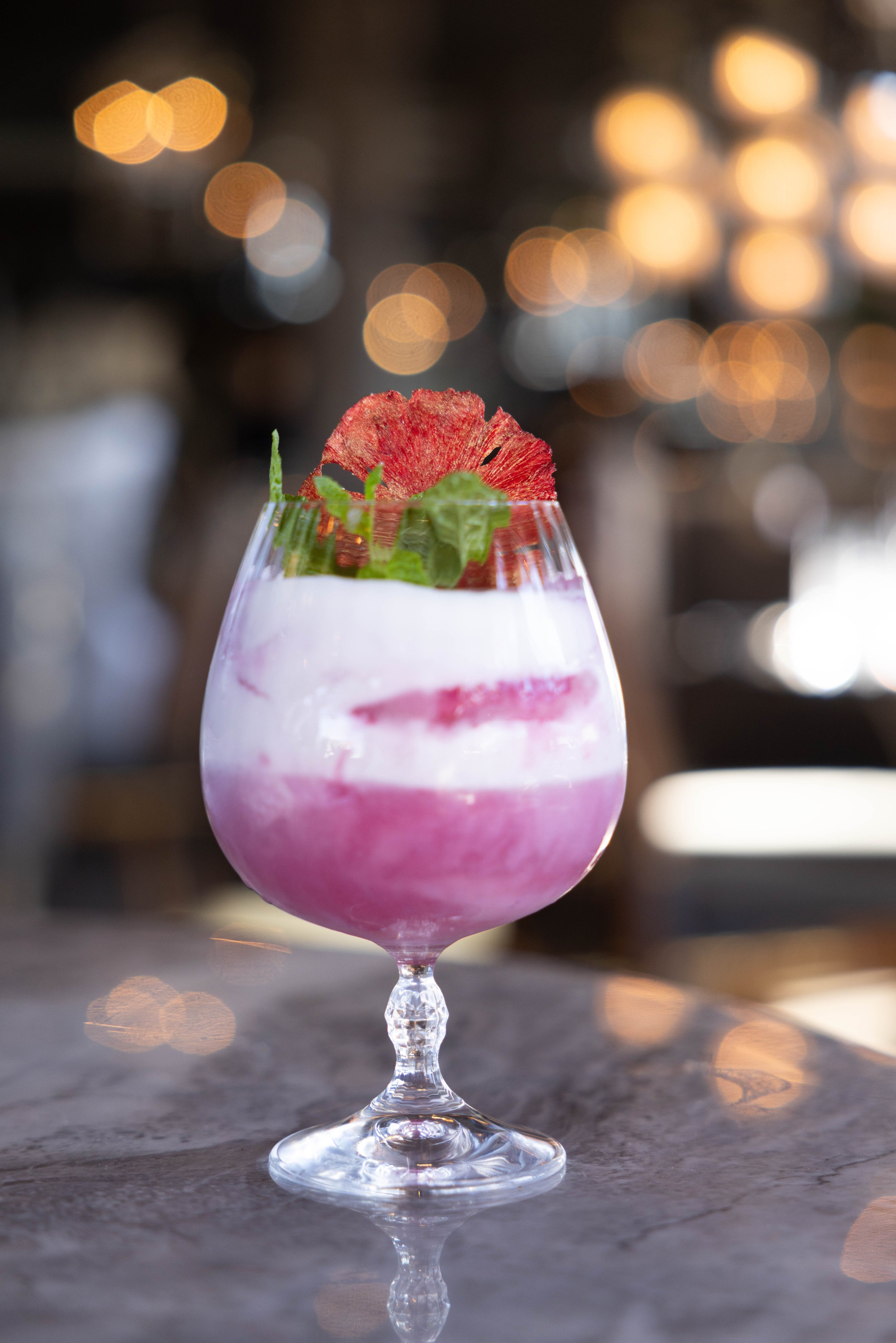 A cocktail in a rounded glass with a short stem, containing pink and white layered drink, garnished with a red flower, green leaf, and shredded vegetable, set on a marble surface with a blurred background of bokeh lights.