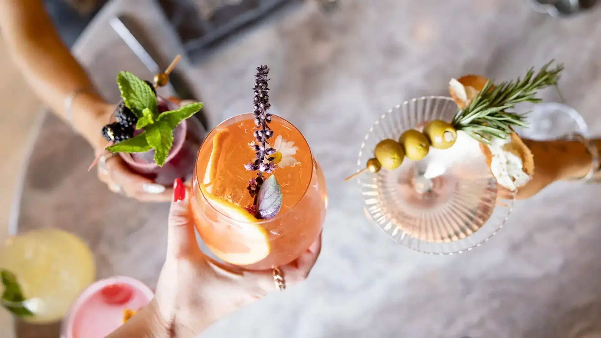 Three colorful cocktails garnished with herbs, fruit, and decorative skewers, viewed from above on a concrete surface.