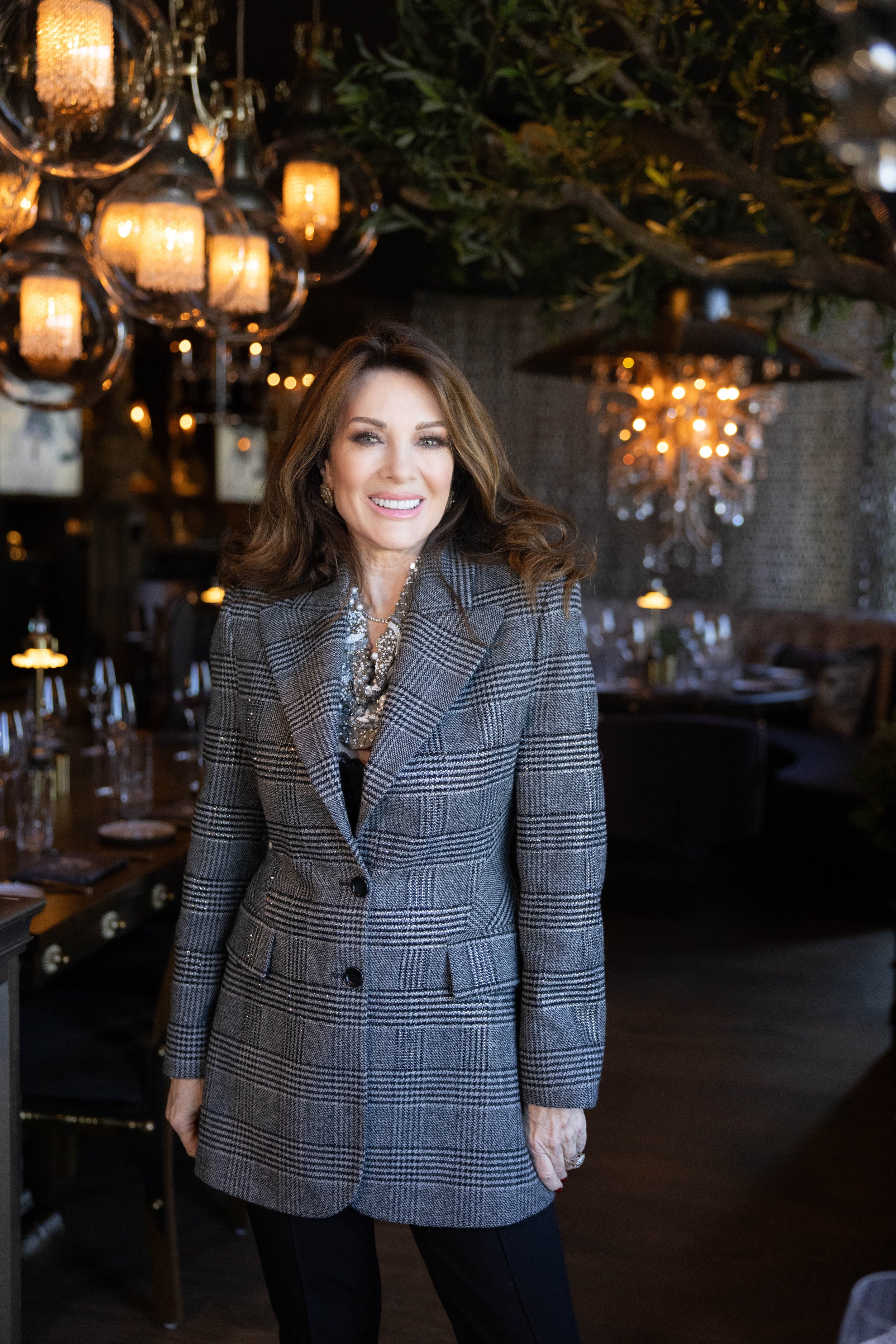 A woman with long brown hair smiling in a stylish gray plaid blazer and black pants, standing in a dimly lit restaurant decorated with chandeliers and warm lighting.