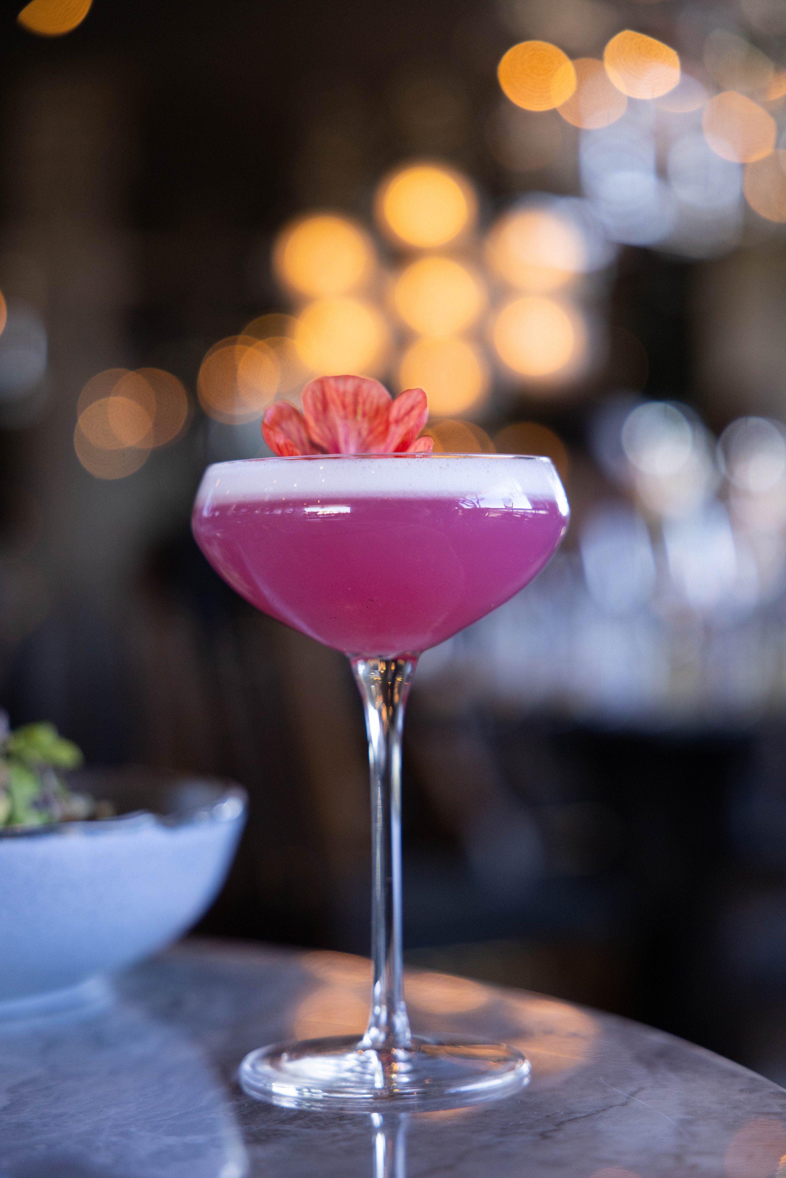 A pink cocktail in a coupe glass garnished with a pink flower on top, set against a background of blurred warm and cool lights.