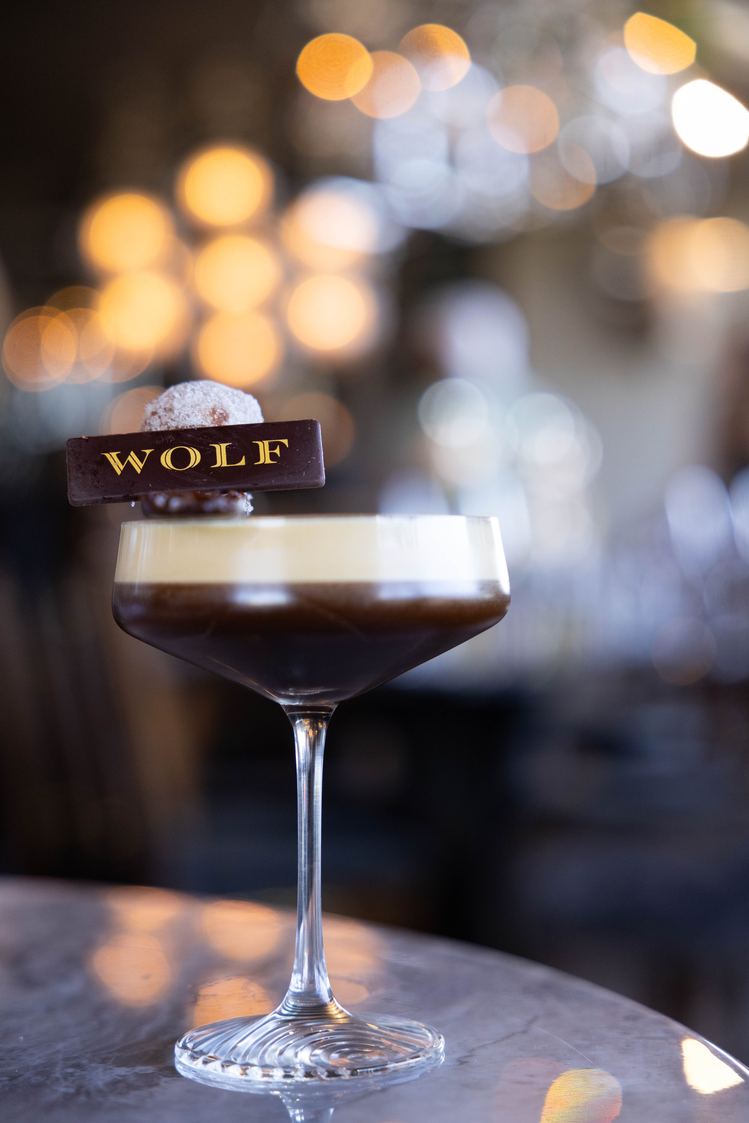 A layered cocktail in a coupe glass with a chocolate garnish. The garnish includes a chocolate piece with the word 'WOLF' written on it and a sugared round. The background is blurred with warm bokeh lights.