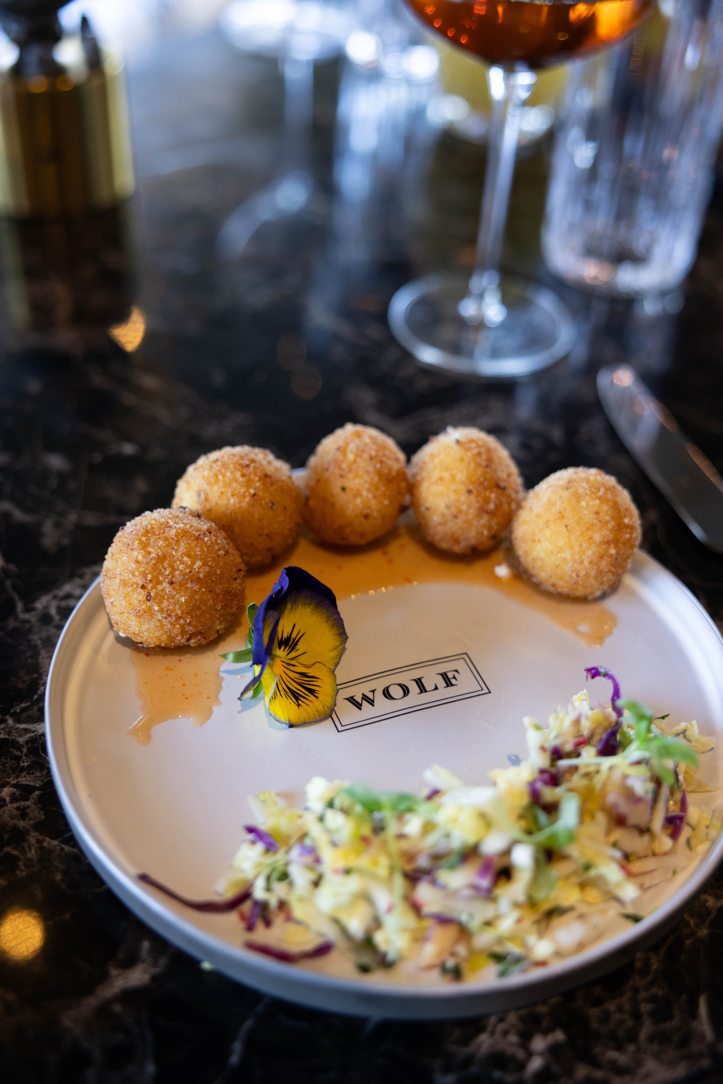 Fried appetizer balls on a plate with a flower garnish and a salad, with wine and water glasses in the background.