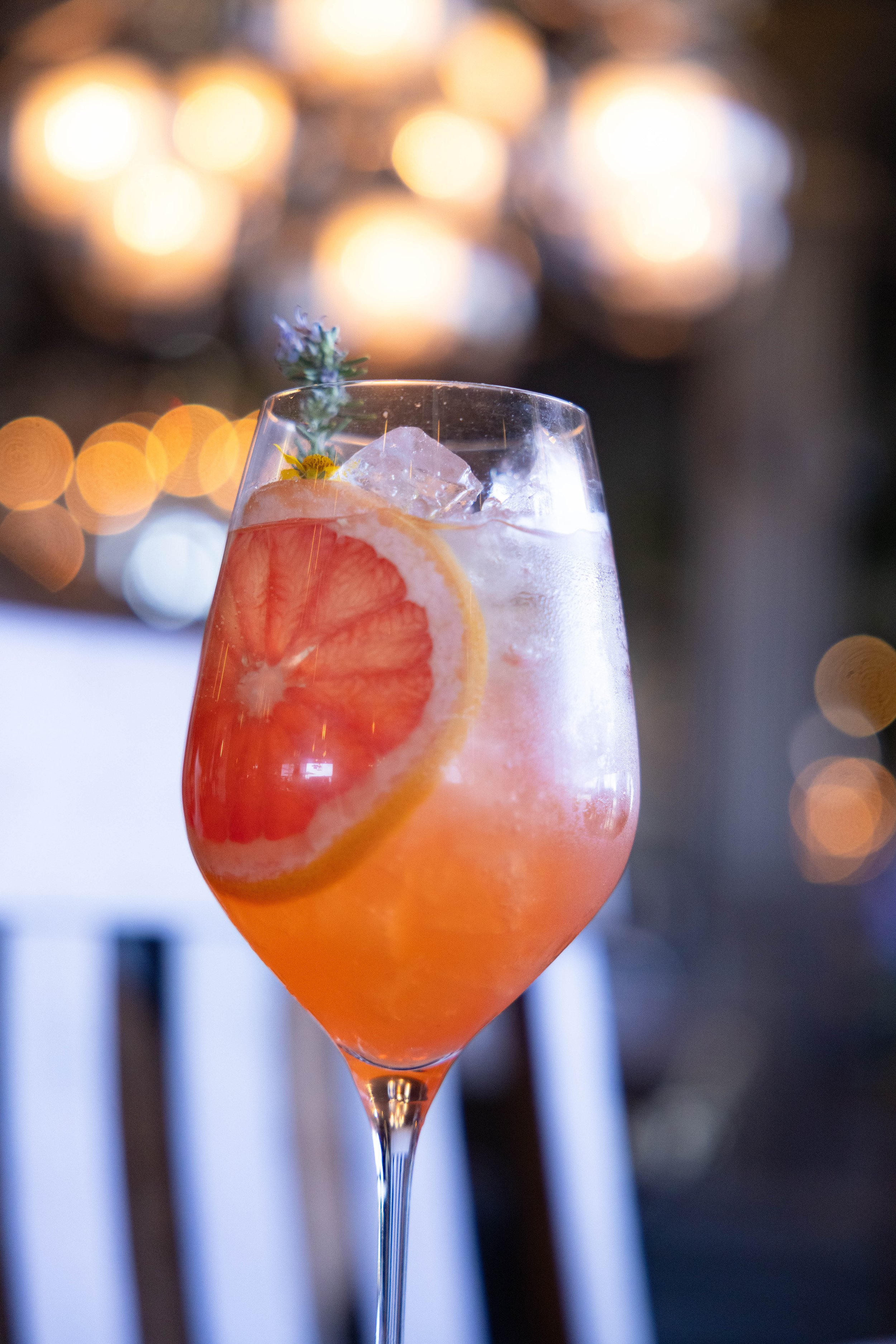 A tall glass of pink cocktail with a slice of grapefruit, ice cubes, and a sprig of lavender, with blurry warm lights in the background.