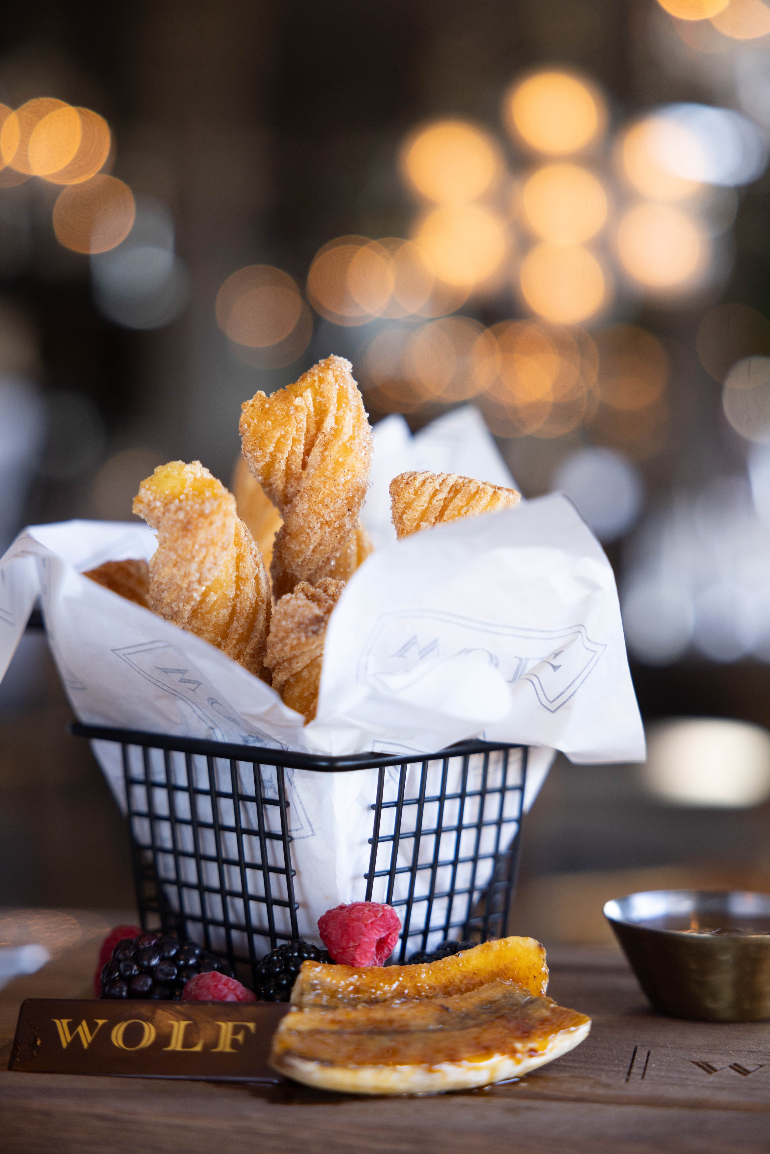 Fried chicken tenders served in a black wire basket with white paper, garnished with berries and a side of sauce on a wooden table with a blurred background of warm lights.