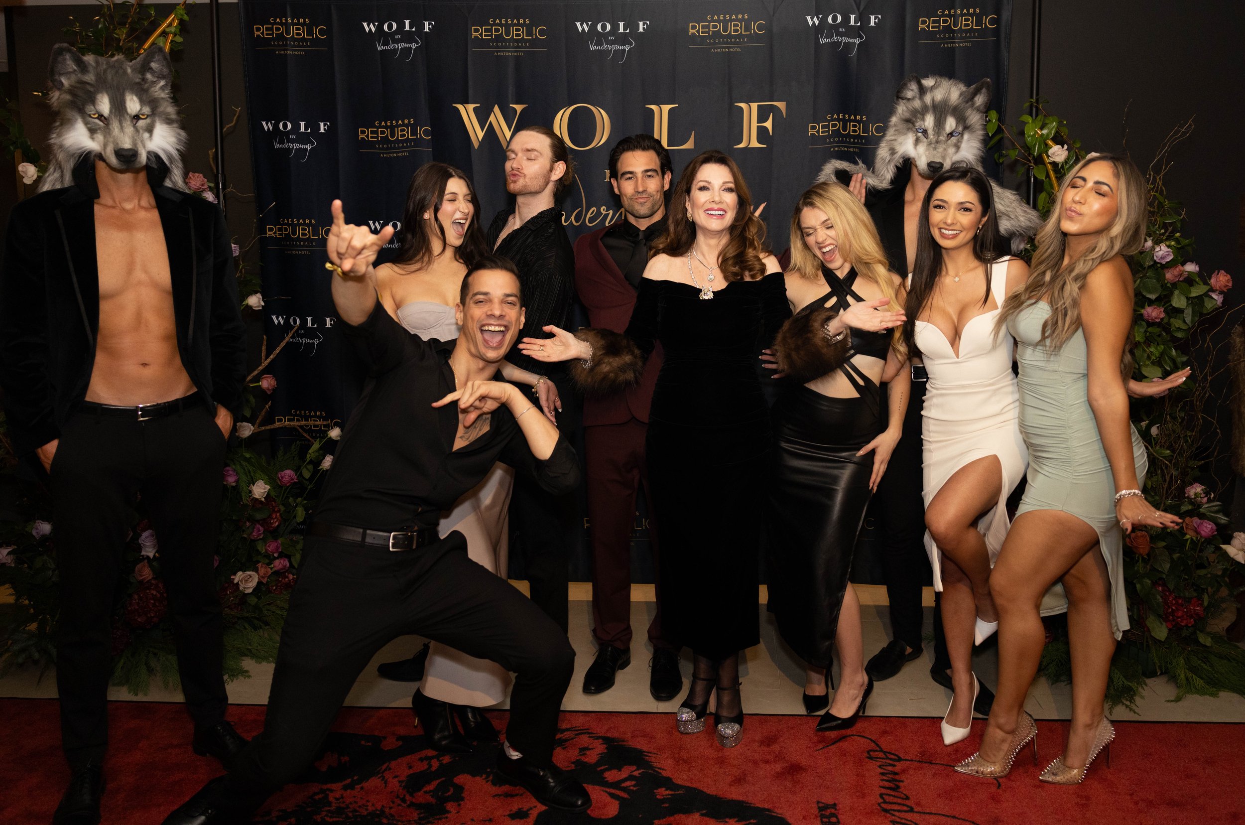 A group of people at a party, some wearing wolf masks, standing in front of a backdrop that reads 'Wolf' and 'Caesars Republic'. They are dressed in formal attire, smiling, posing, and making celebratory gestures.