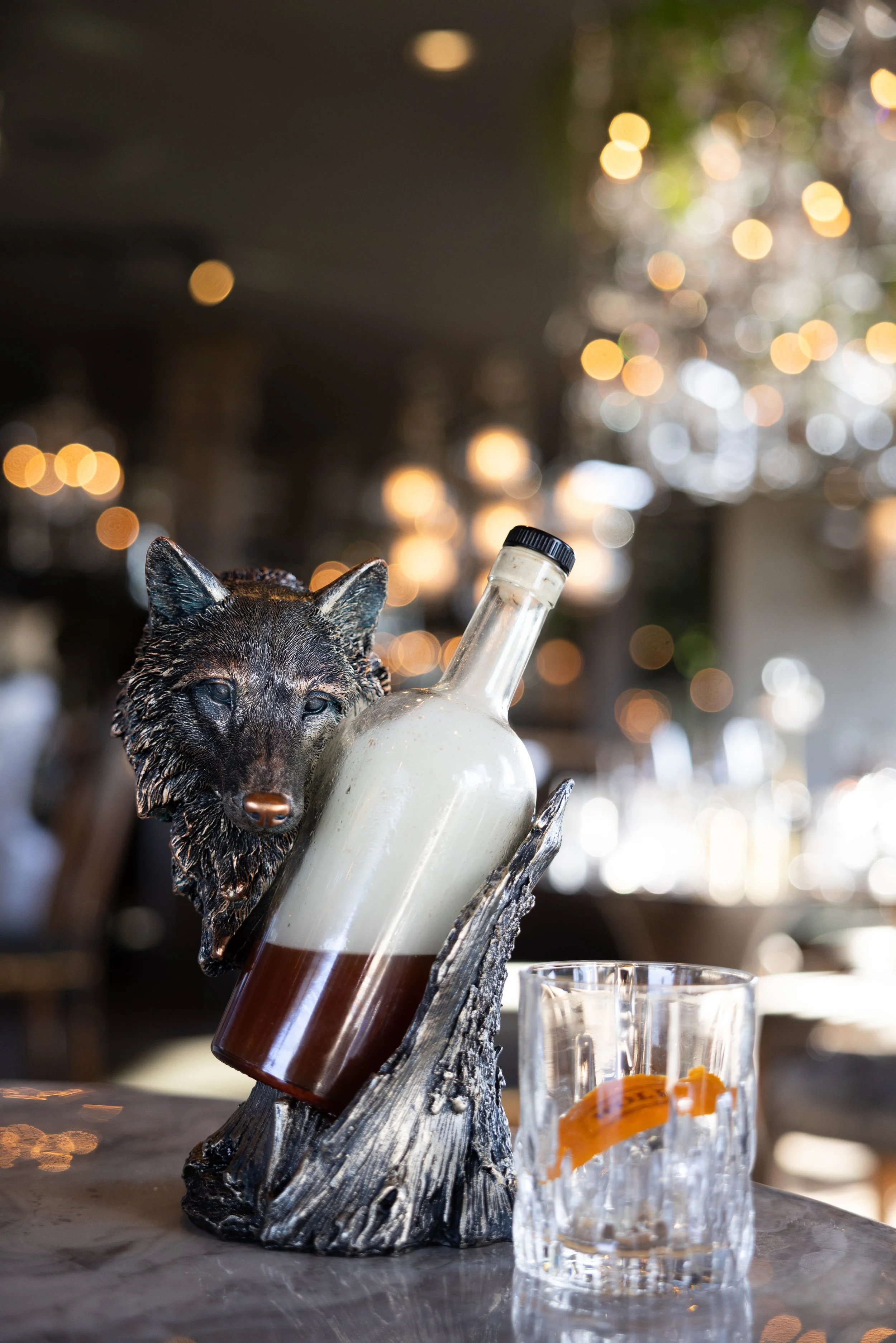 A decorative bottle with a wolf head sculpture is placed in a wooden holder, with a glass of ice and orange peel to the side, in a restaurant setting with warm lighting and bokeh background.